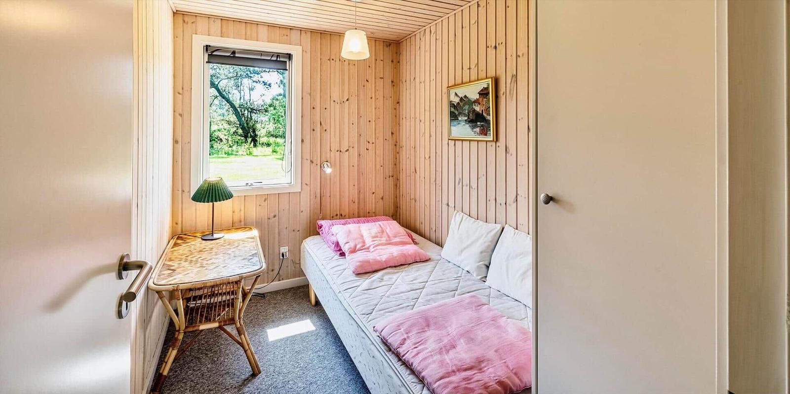 Schlafzimmer mit Holzwand, Bett, Tisch und Fenster zur grünen Umgebung.