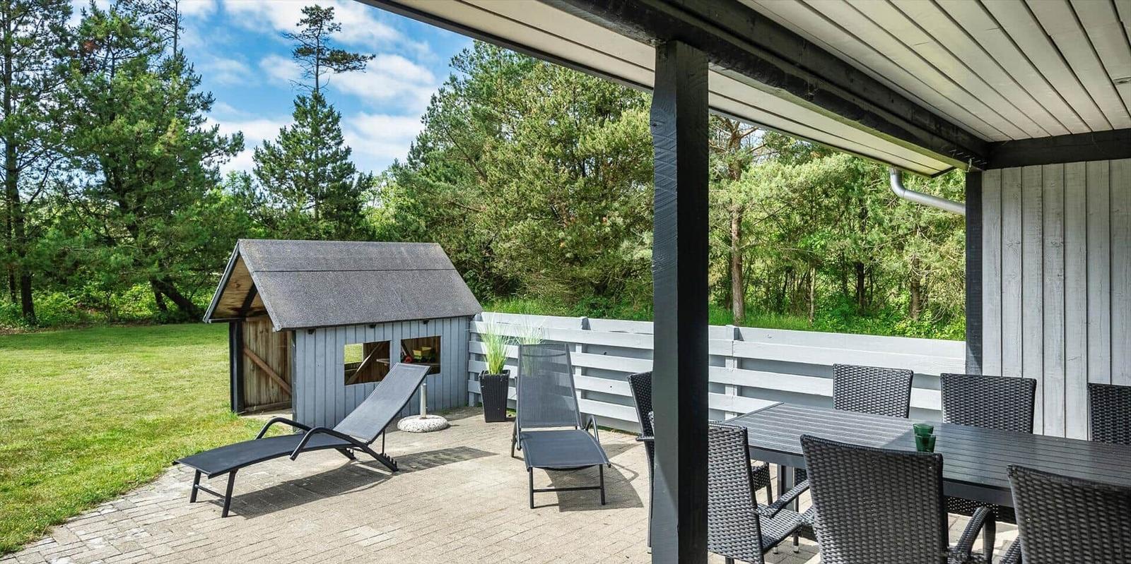 Terrasse mit Tisch, Stühlen und Liegestühlen. Im Hintergrund ein kleines Holzhaus und Wald.
