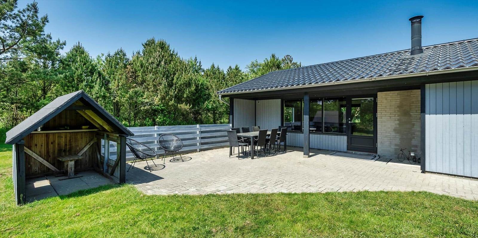 Haus mit Terrasse, Tisch, Stühlen und Grillhütte im Grünen.