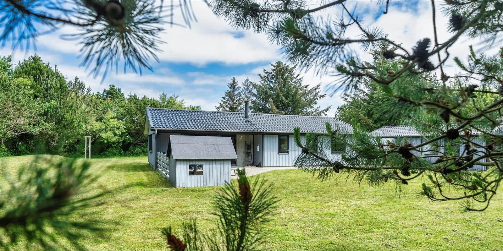 Ein Haus mit grauem Dach und weißem Anbau steht auf grünem Rasen. Umgeben von Bäumen und Nadelbäumen.