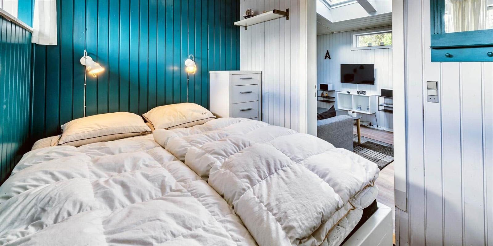 Schlafzimmer mit Bett, blauer Wand und Blick in Wohnbereich mit TV.