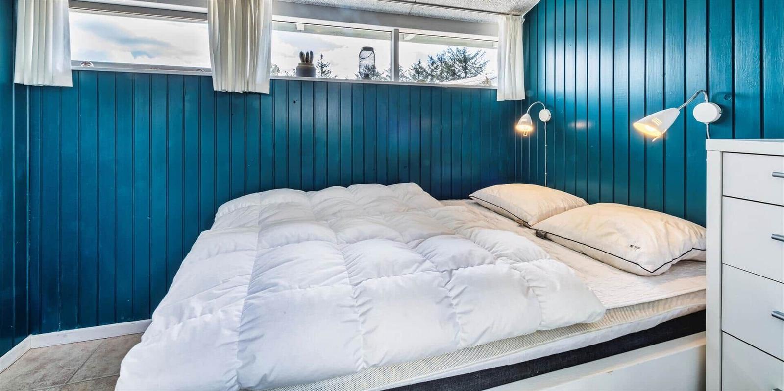 Schlafzimmer mit blauen Wänden, Bett mit weißer Bettwäsche und Fenster mit Blick auf Bäume.