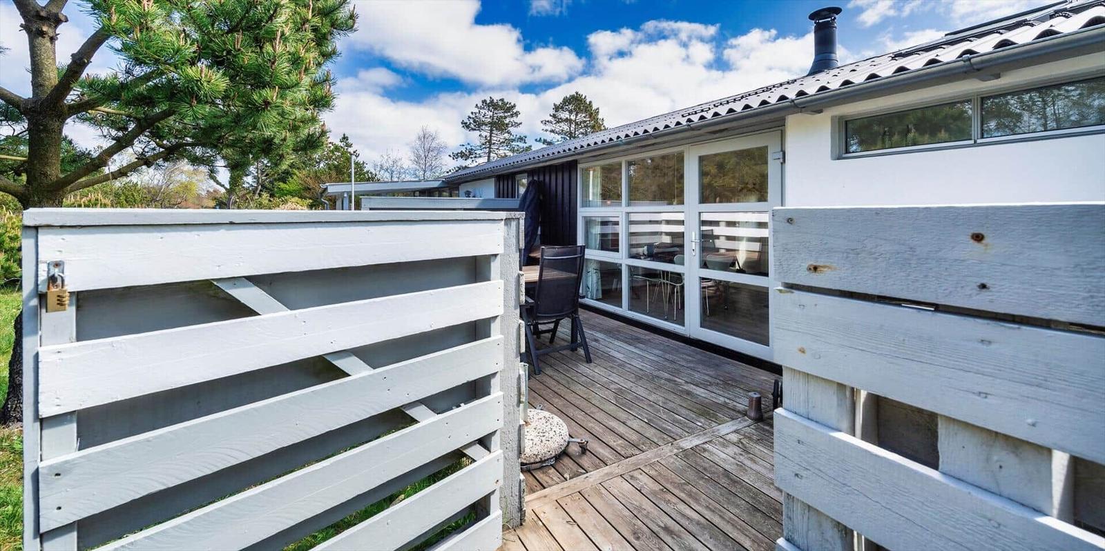 Terrasse mit Sitzgelegenheit und Glasfront zum Haus. Holzgeländer und grüne Umgebung.