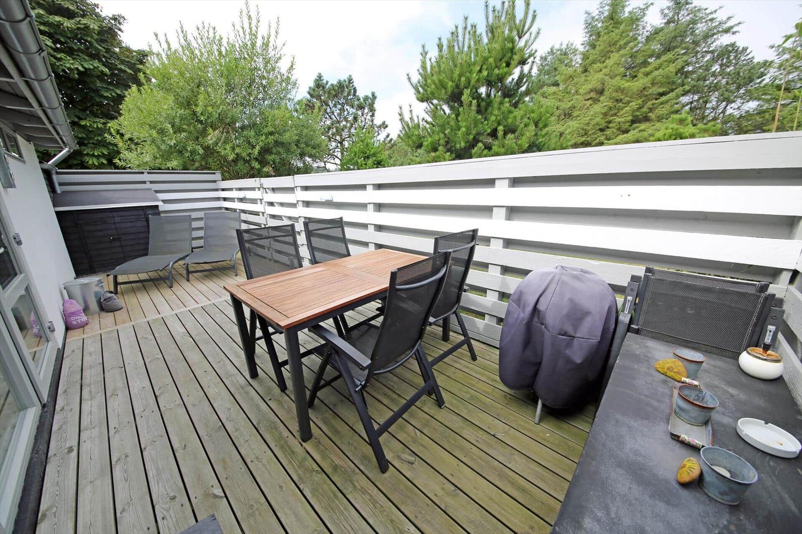 Terrasse mit Tisch, Stühlen, Grill und Liegestühlen. Hintergrund: Holzgeländer und Bäume.