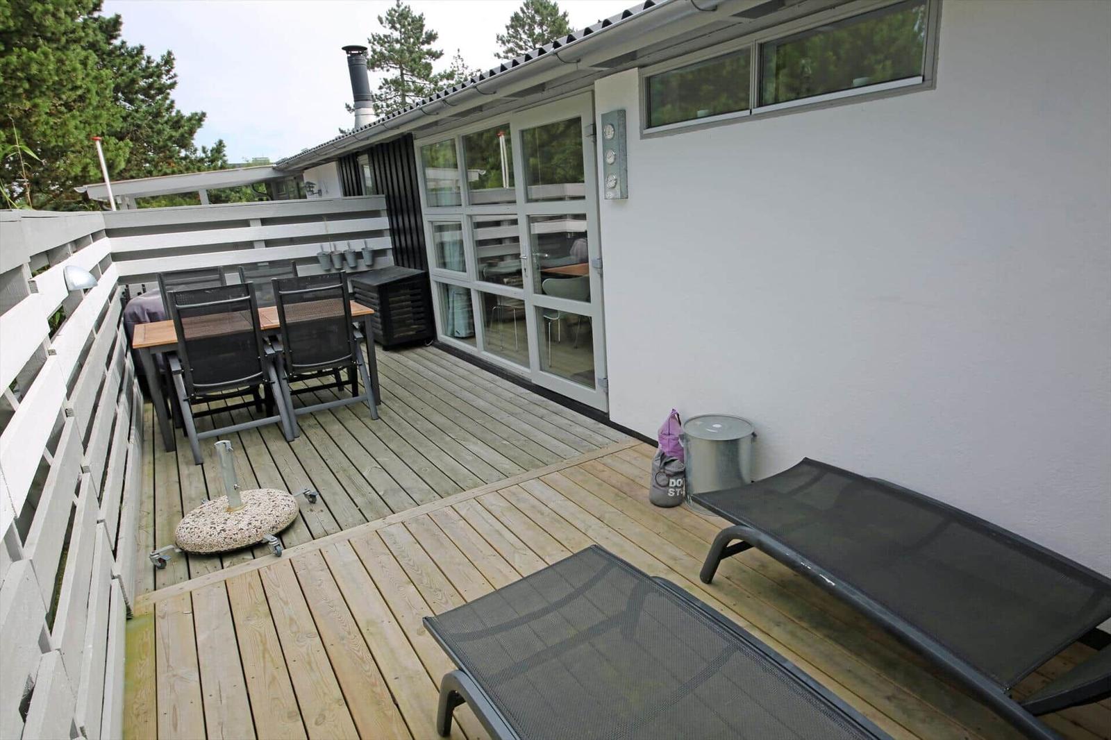 Terrasse mit Tisch, Stühlen und Liegen. Hintergrund: Haus mit Glasfront und Bäumen.