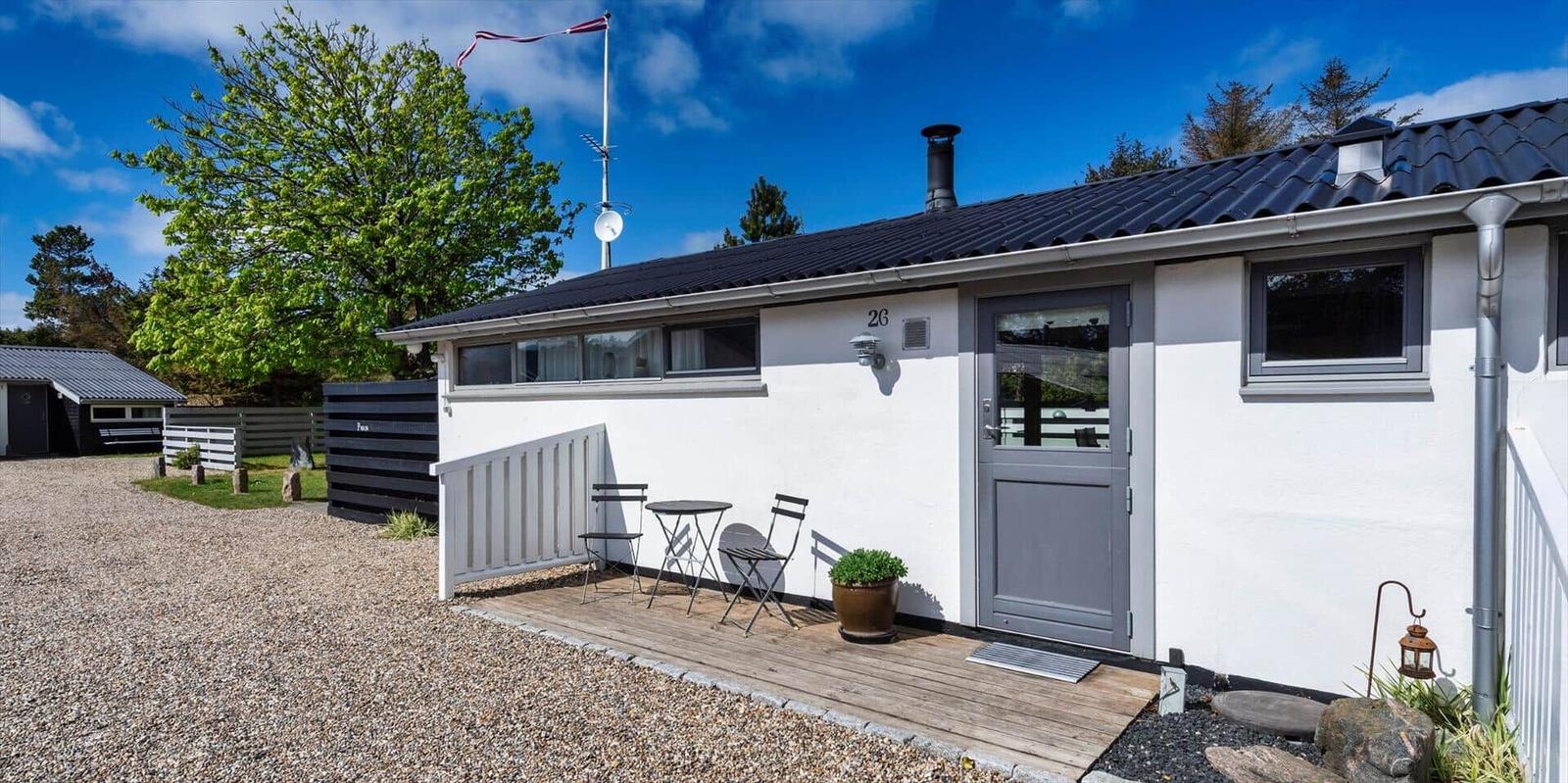 Wohnhaus mit grauer Tür, Holzterrasse und Gartenmöbeln. Schwarzer Dachfirst und weiße Außenwände.