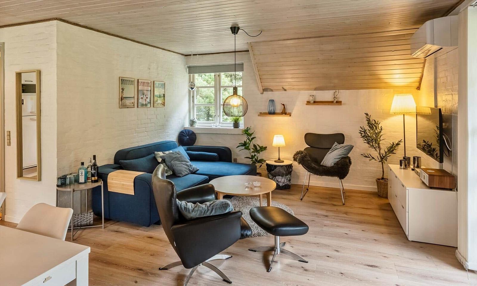 Wohnzimmer mit blauem Sofa, schwarzen Sesseln und Holzboden. Fenster, Pflanzen und Beleuchtung sichtbar.