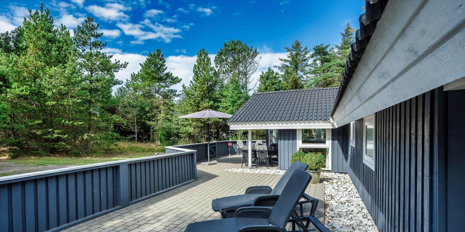 Terrasse mit Liegestühlen und Sitzbereich unter einem Sonnenschirm. Hintergrund: Wald und blauer Himmel.
