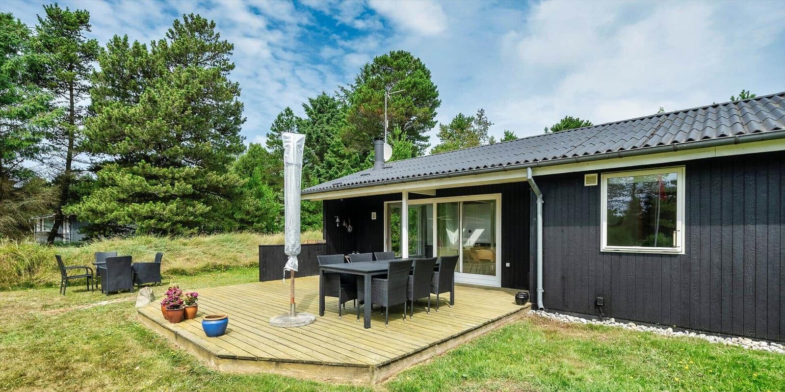 Schwarzes Holzhaus mit Terrasse und Garten. Sichtbar: Tisch, Stühle, Sonnenschirm, Fenster und Bäume.
