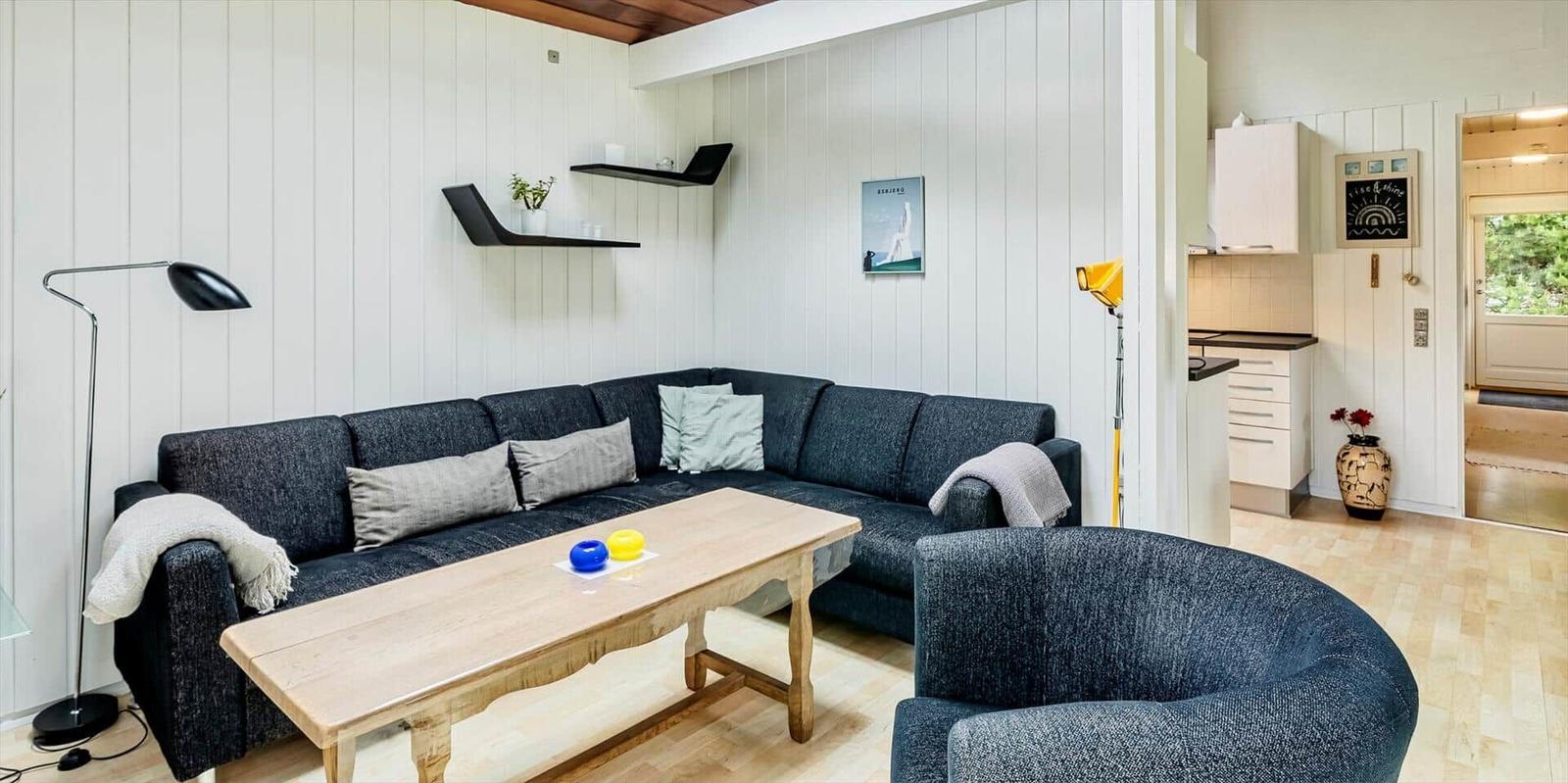 Wohnzimmer mit Sofa, Tisch und Sessel. Küche und Eingangsbereich sichtbar.