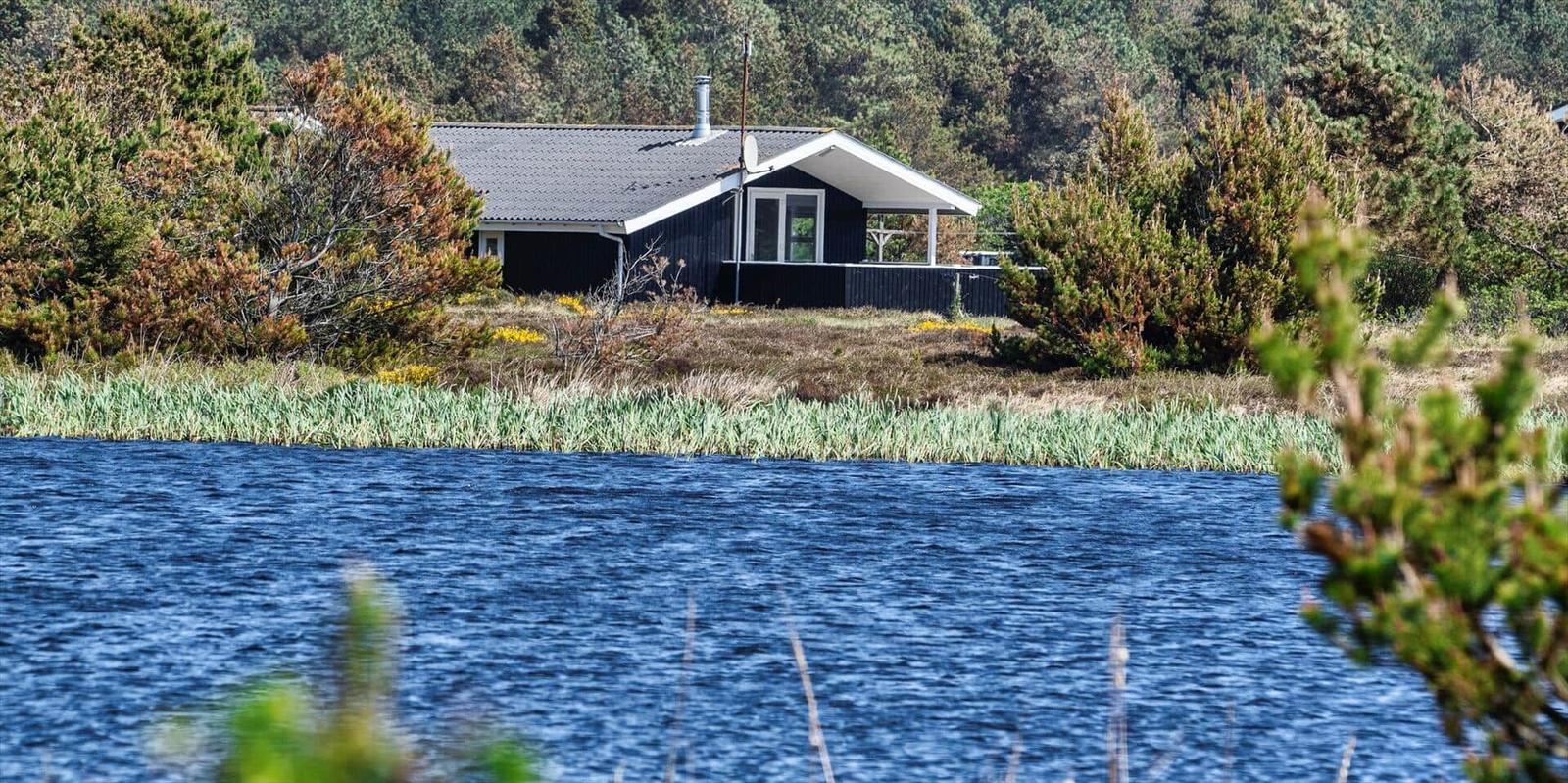 Ein dunkelblau gestrichenes Haus mit weißem Dachrand steht am Ufer eines Sees, umgeben von Bäumen und Gras.