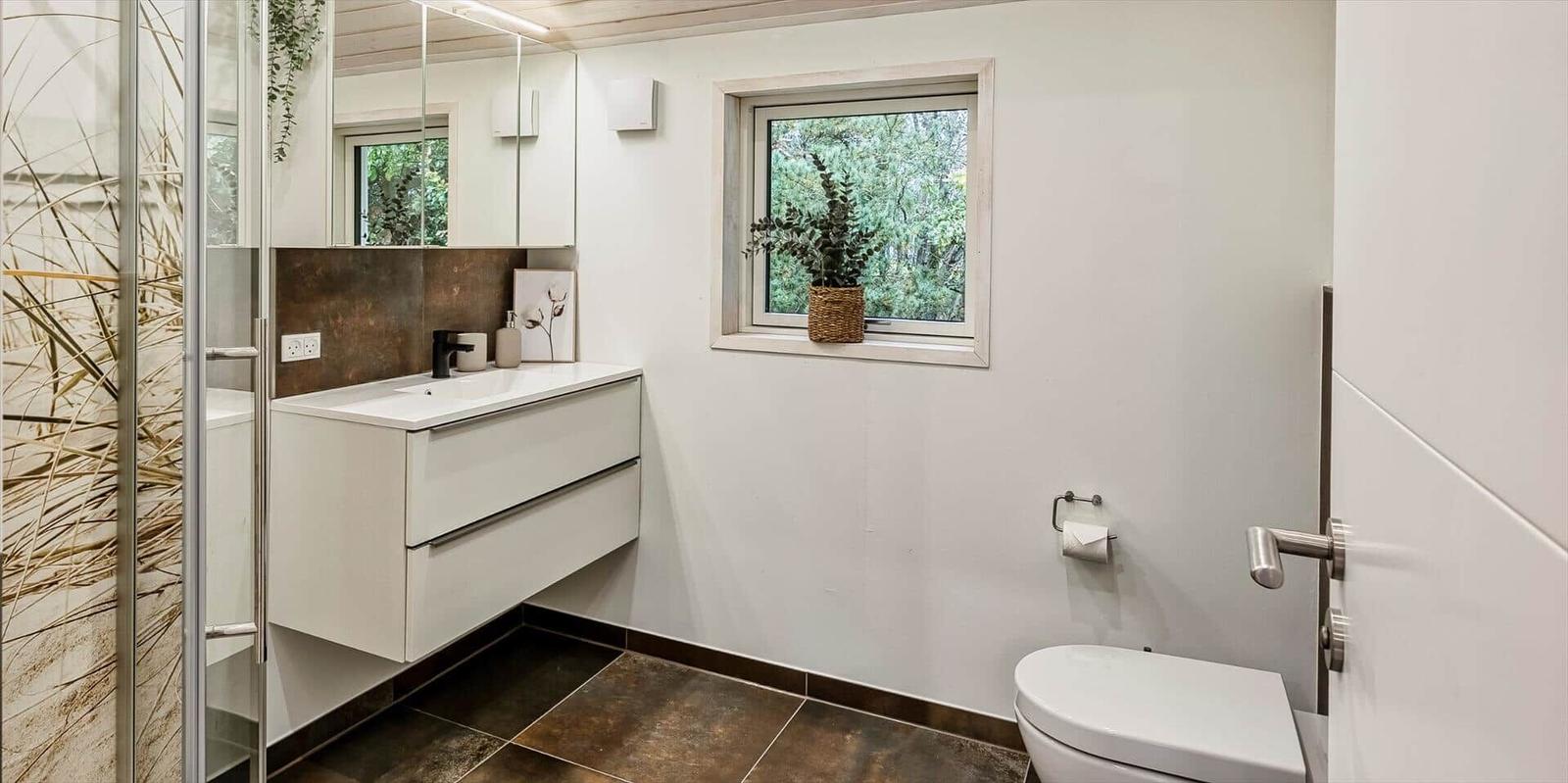 Badezimmer mit Dusche, Waschbecken und Toilette. Fenster mit Pflanze.