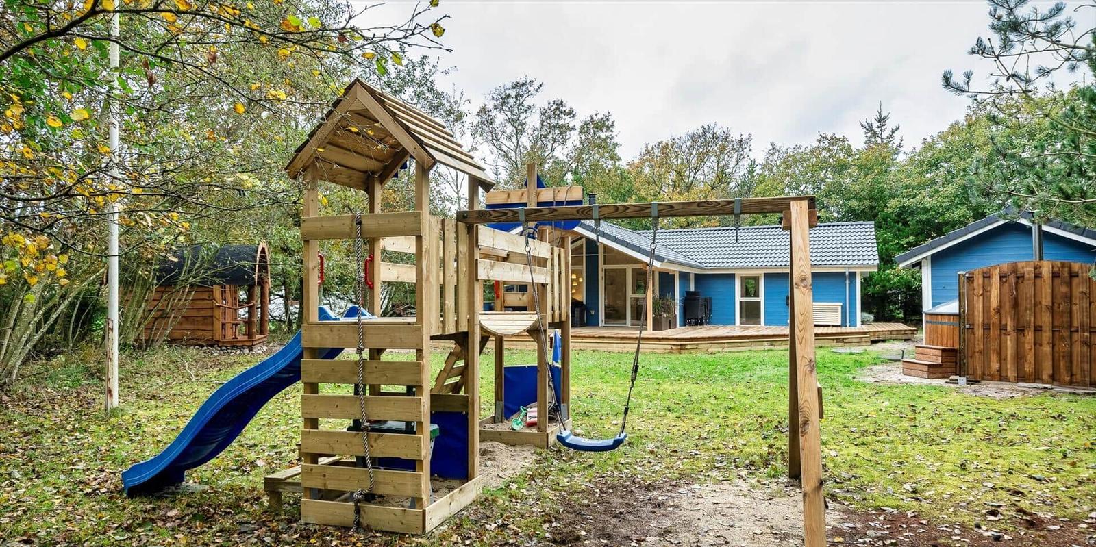 Holzspielplatz mit Rutsche und Schaukel vor einem blauen Haus mit Terrasse.