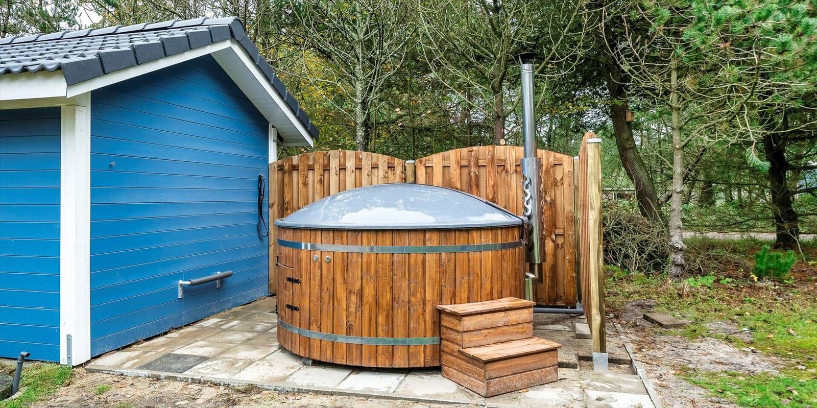 Holzwhirlpool mit Metallrahmen neben blauem Gebäude im Wald.