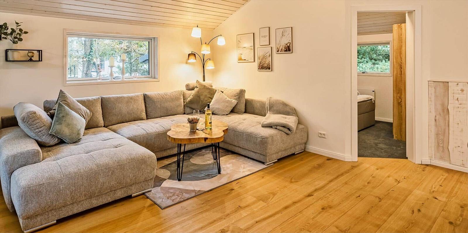 Wohnzimmer mit Ecksofa, Holzboden und Tisch. Durchgang zu Schlafzimmer.
