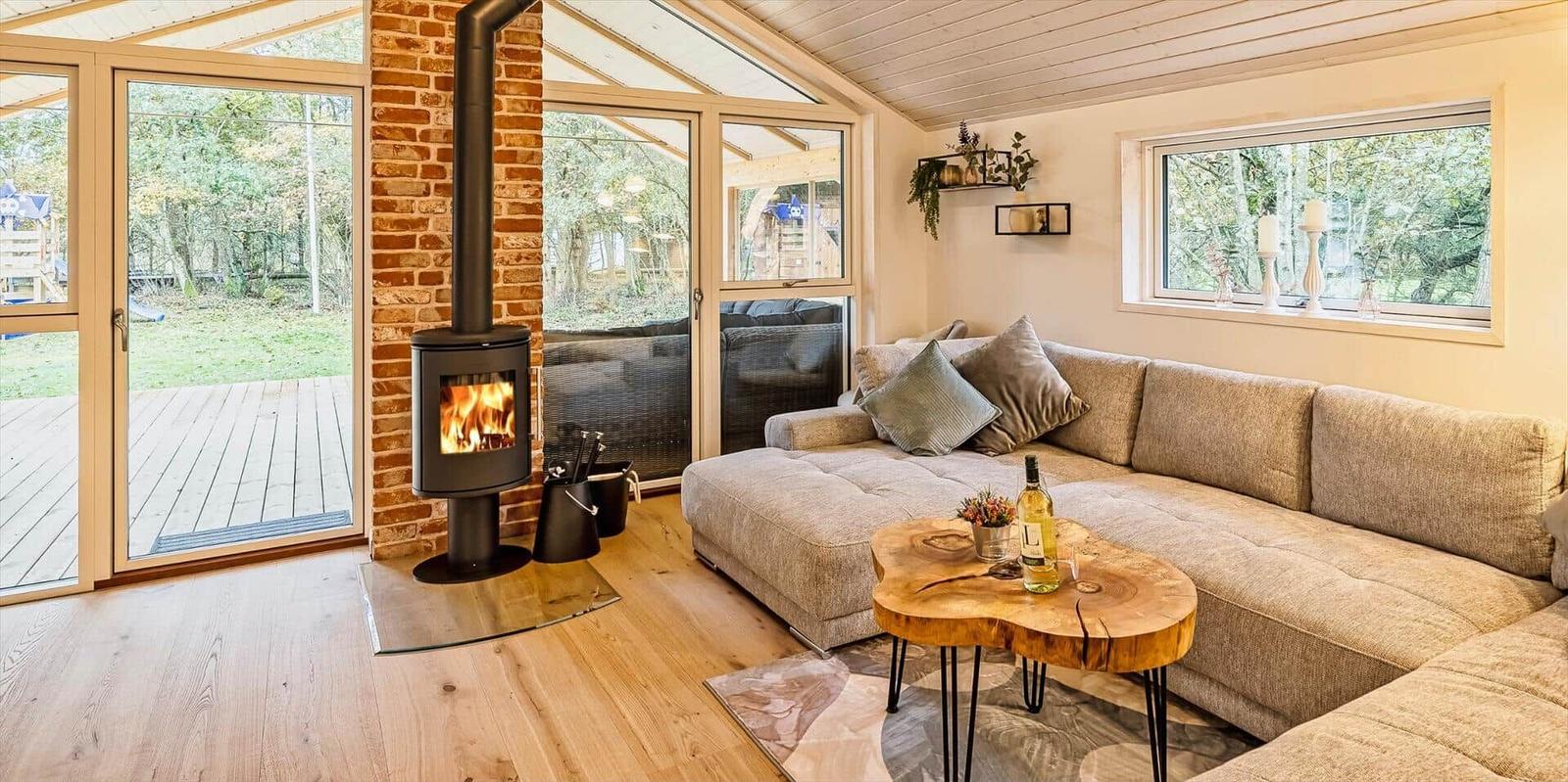 Wohnzimmer mit Kamin, Sofa und Holztisch. Große Fenster und Tür zur Terrasse.