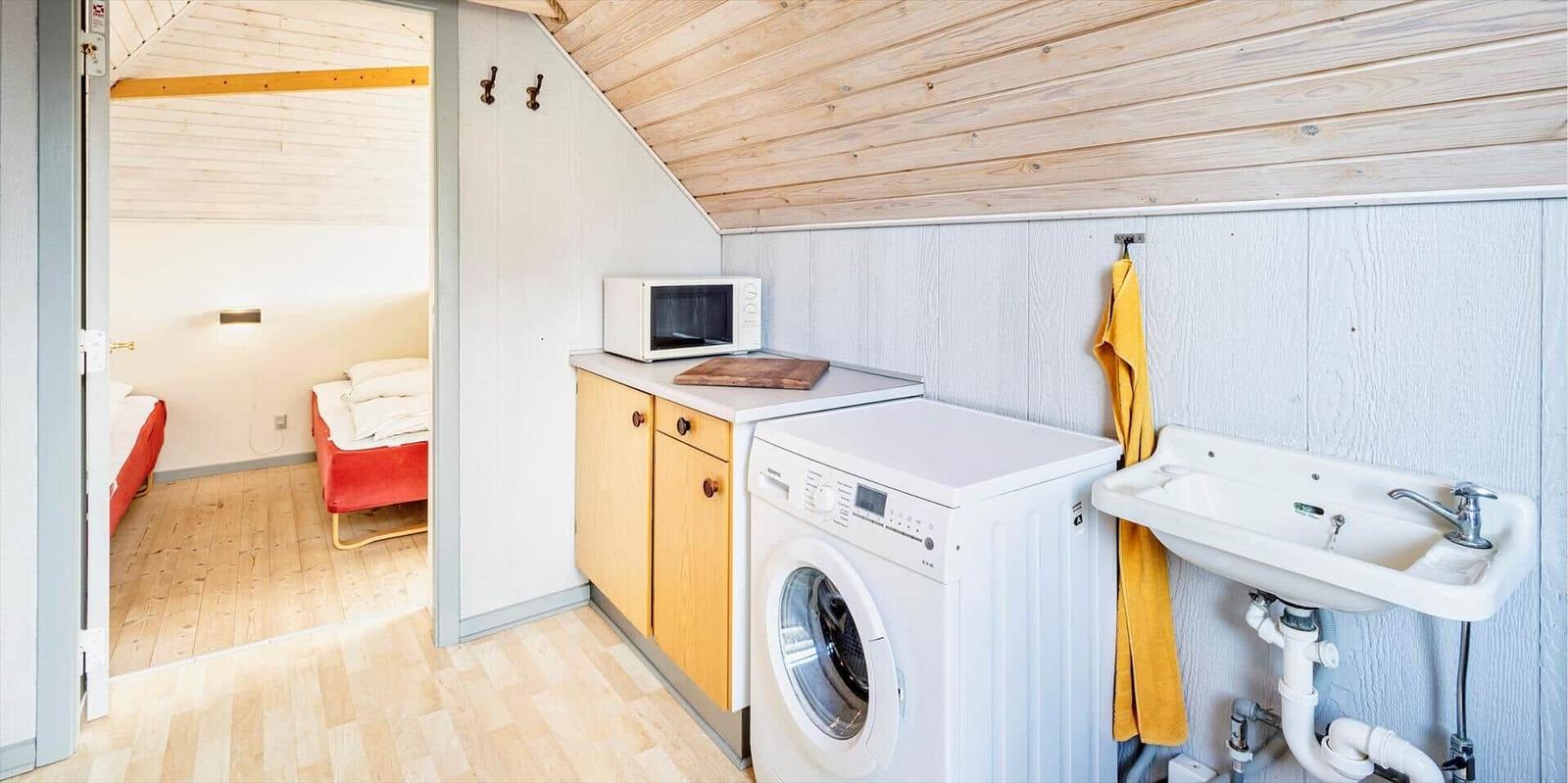 Wohnbereich mit Waschmaschine, Spüle und Mikrowelle. Im Hintergrund ist ein Schlafzimmer mit rotem Bett zu sehen.