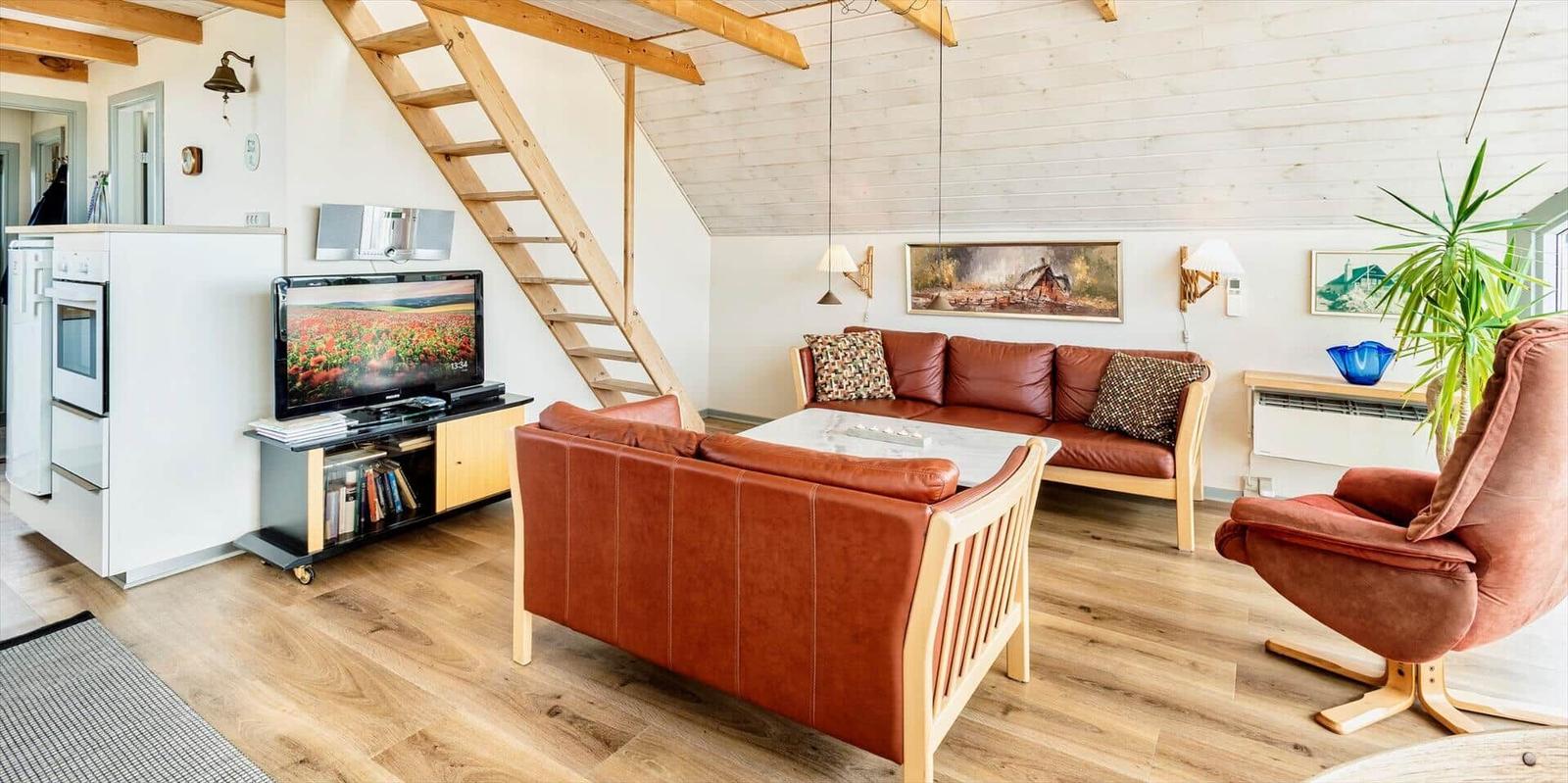Wohnzimmer mit Sofa, Sessel und TV. Holzdecke und Treppenhaus sichtbar.