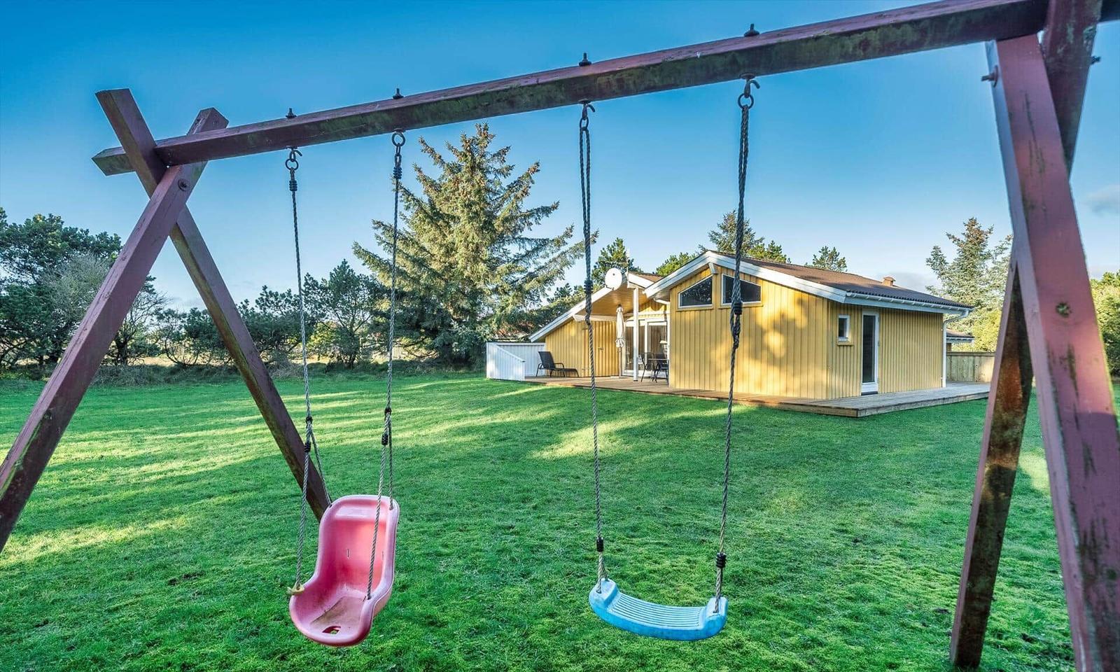 Ein gelbes Ferienhaus mit Balkon steht auf einer grünen Wiese hinter einem Schaukelgestell.