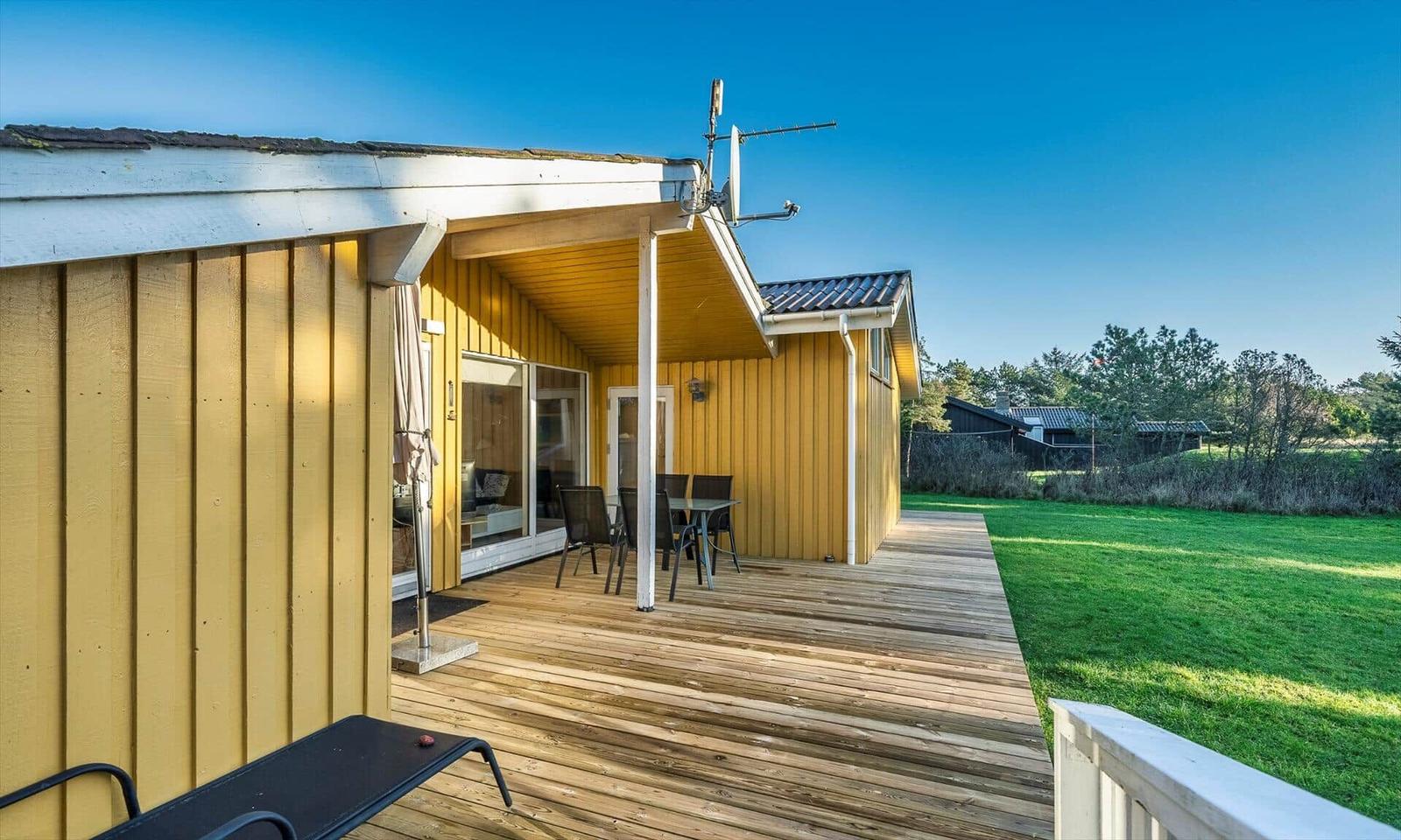 Gelbes Ferienhaus mit Holzterrasse und Garten. Tisch und Stühle sind draußen.