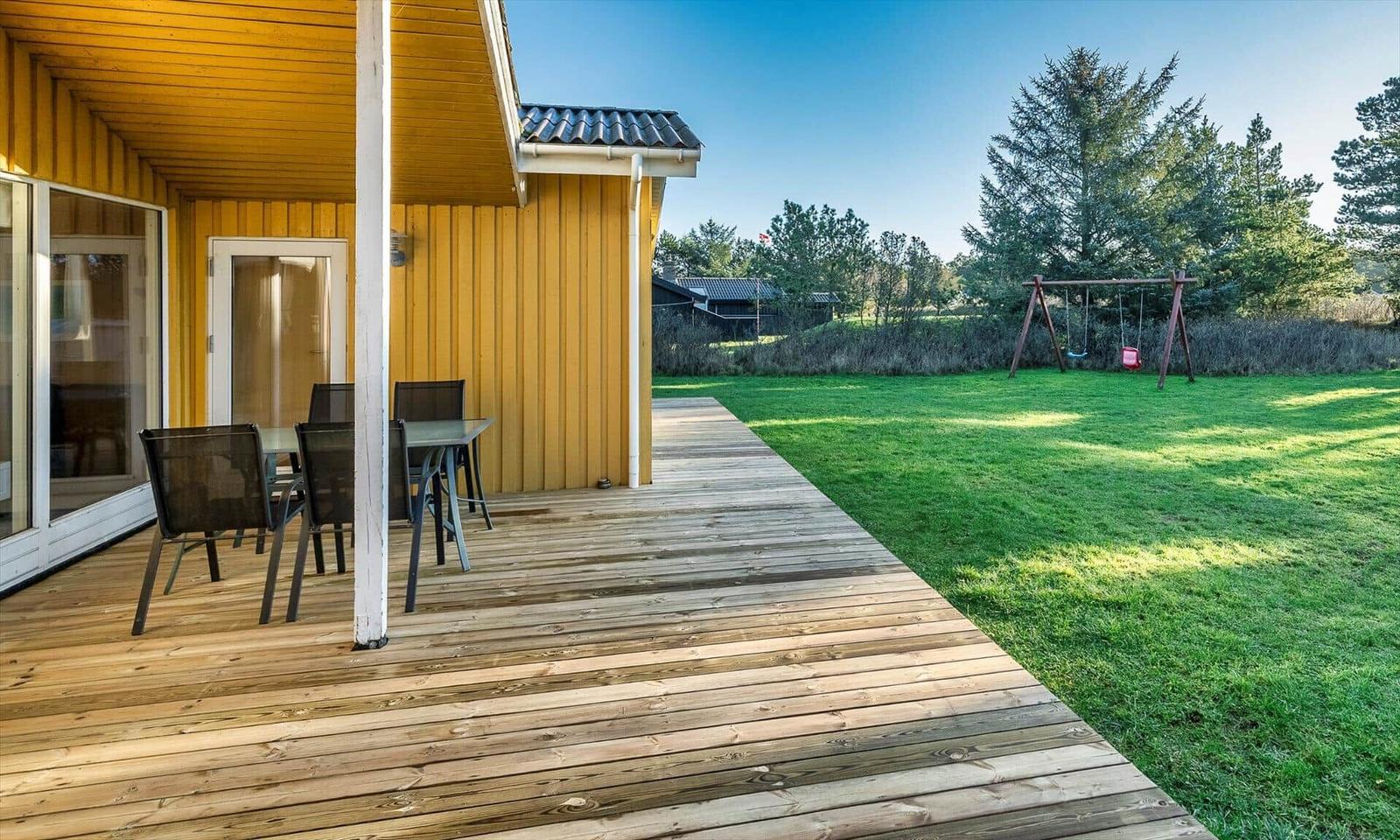 Holzterrasse mit Tisch und Stühlen neben gelbem Haus. Grasfläche mit Schaukel im Hintergrund.