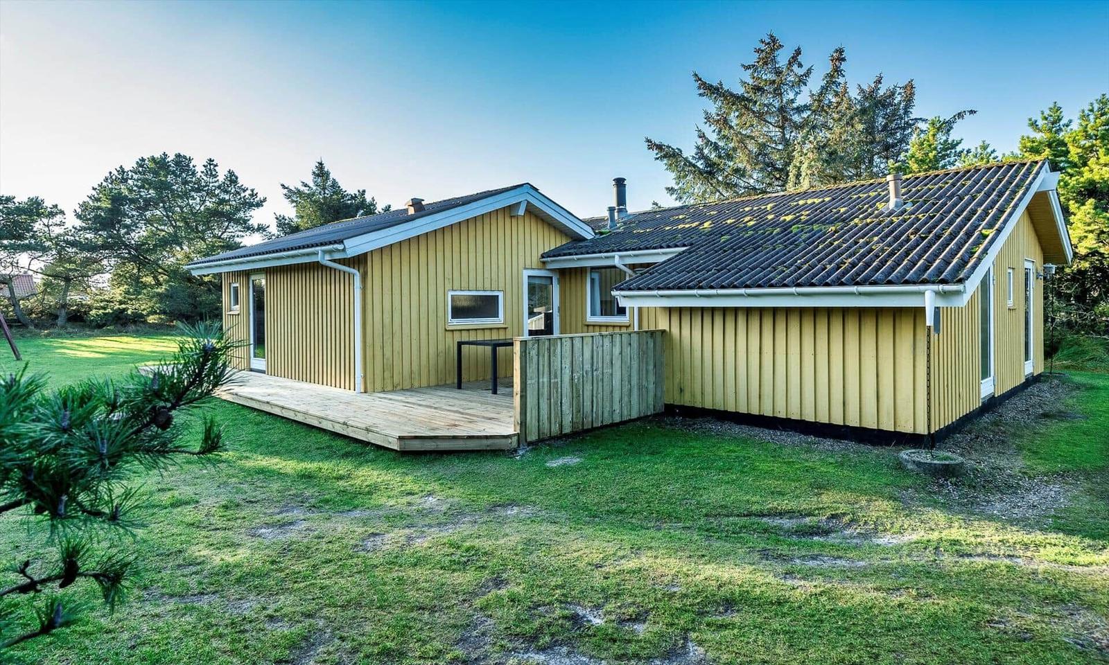 Ein gelbes Ferienhaus mit Holzveranda und Garten. Umgeben von Bäumen und Gras.