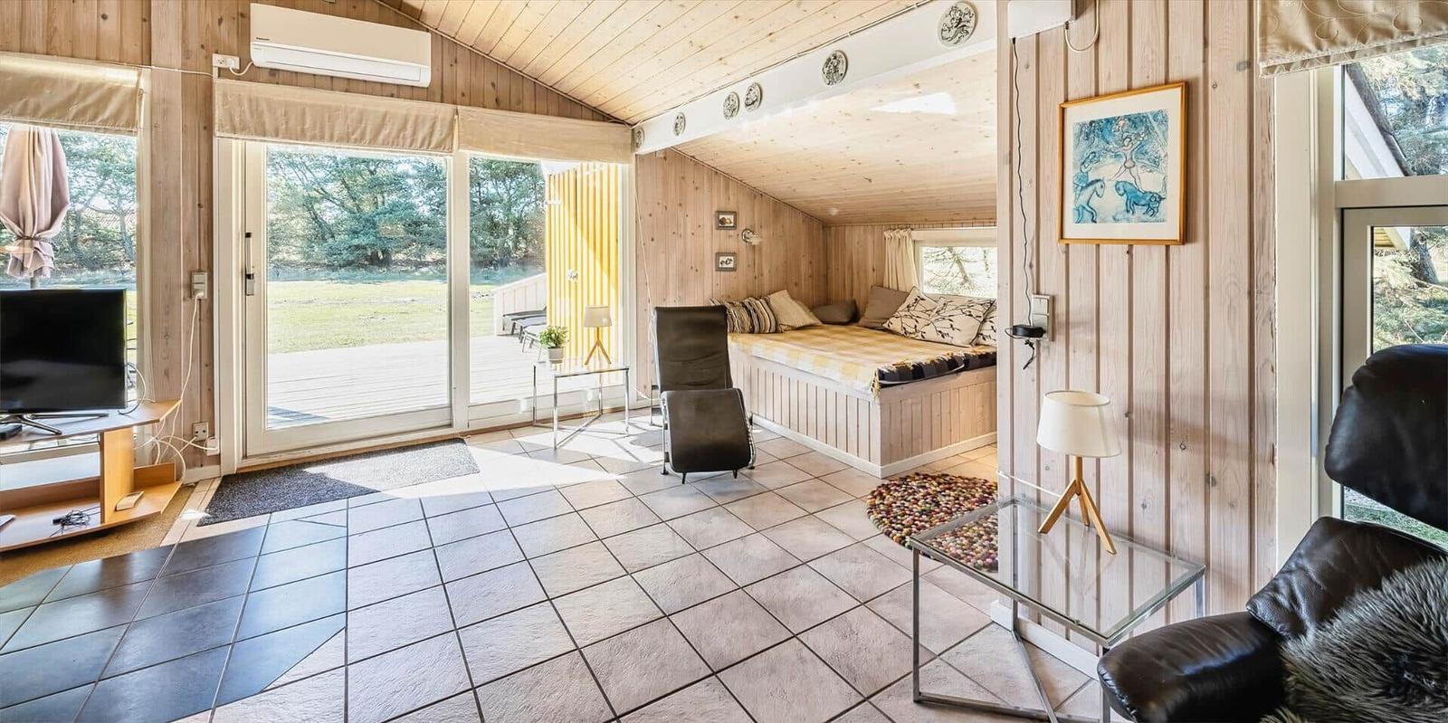 Wohnraum mit Schlafbereich, Holzwand und Terrassentür. Fliesenboden und Sitzmöbel.