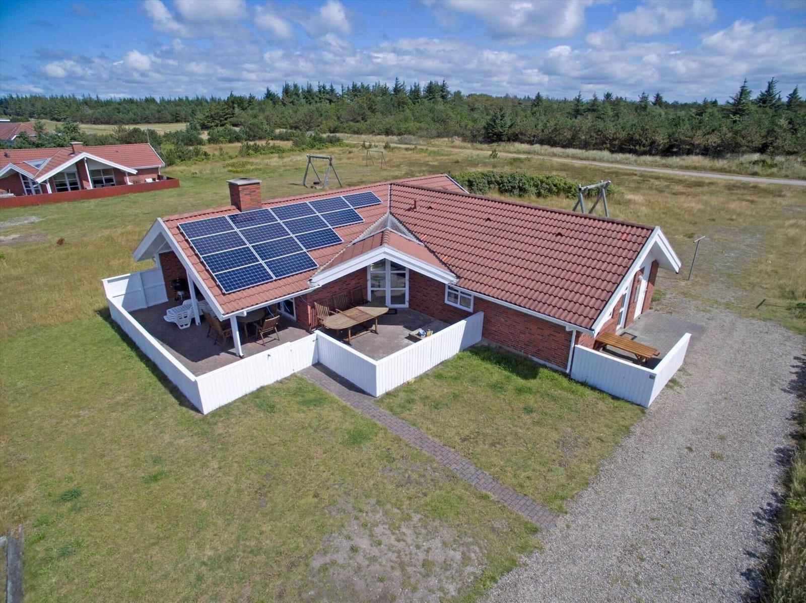Rotes Dach mit Solarpaneelen, weiße Umzäunung, Grillplatz und Sitzgelegenheiten im Garten.