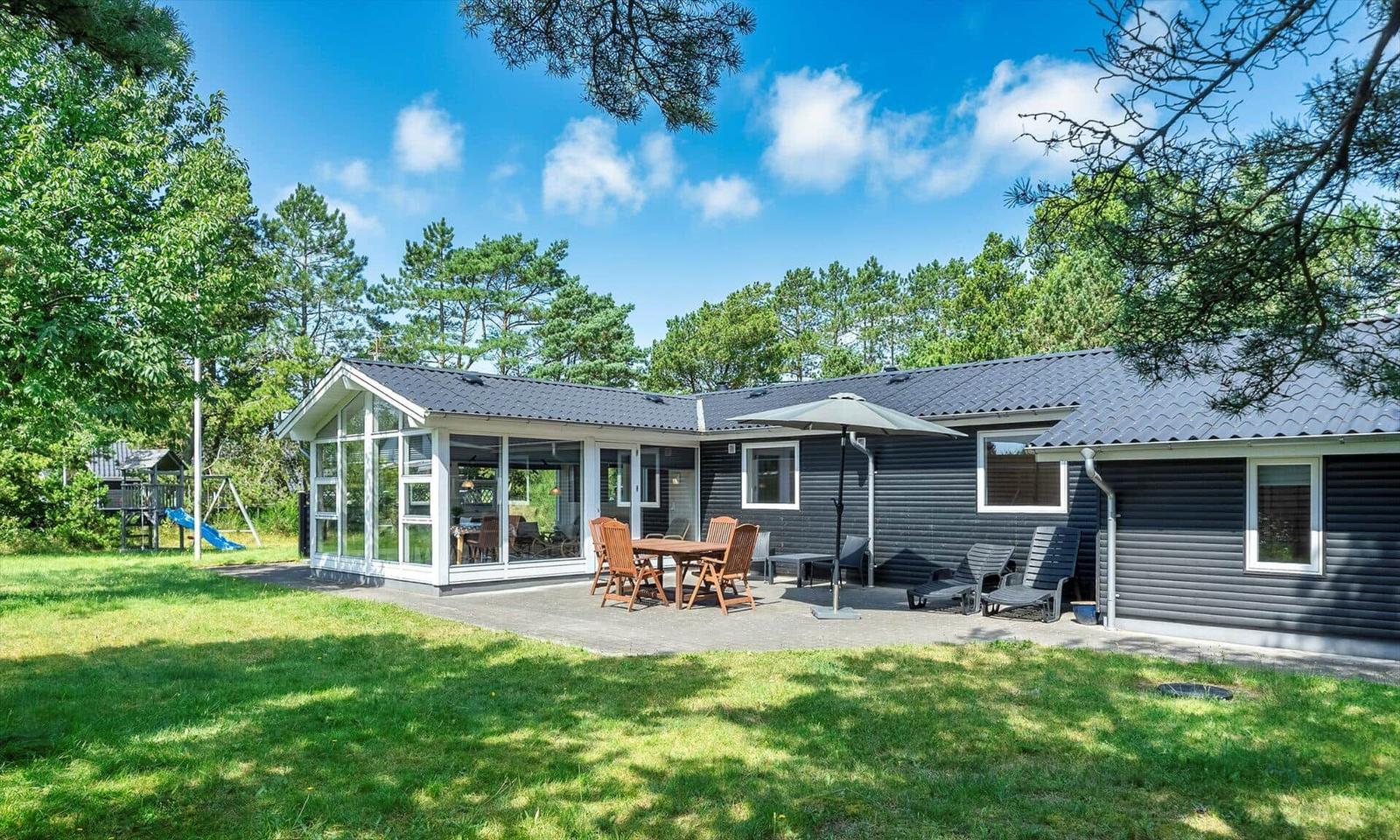 Moderne Ferienhaus mit Terrasse, Garten und Spielplatz unter Bäumen.