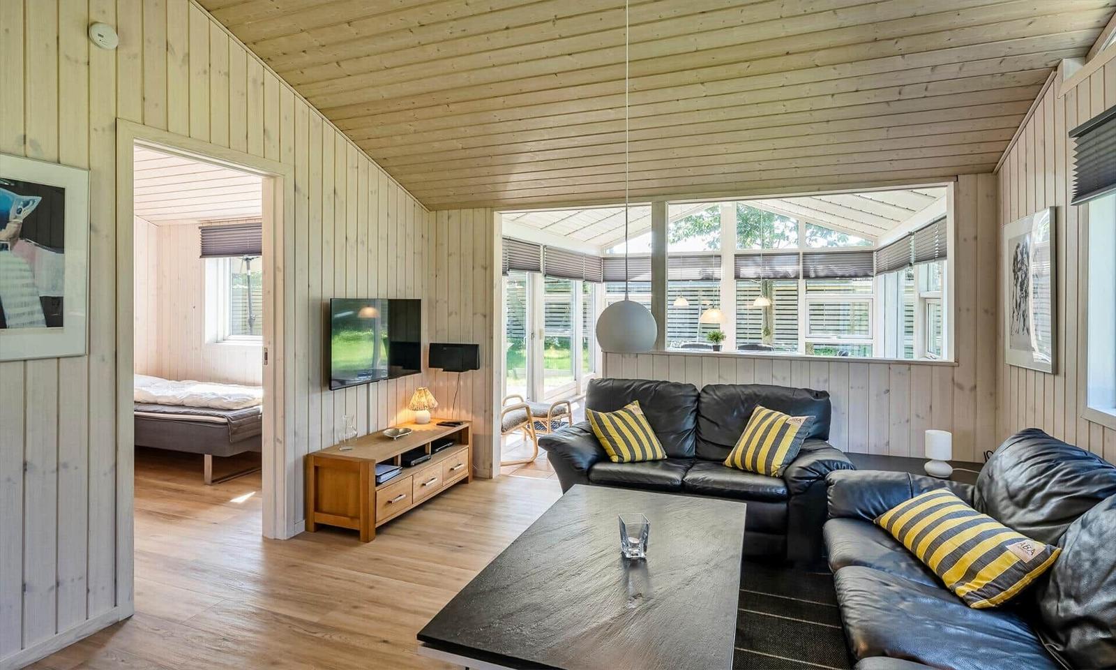 Wohnzimmer mit Ledersofa, Holzwand und TV. Durchgang zur Schlafzimmer.
