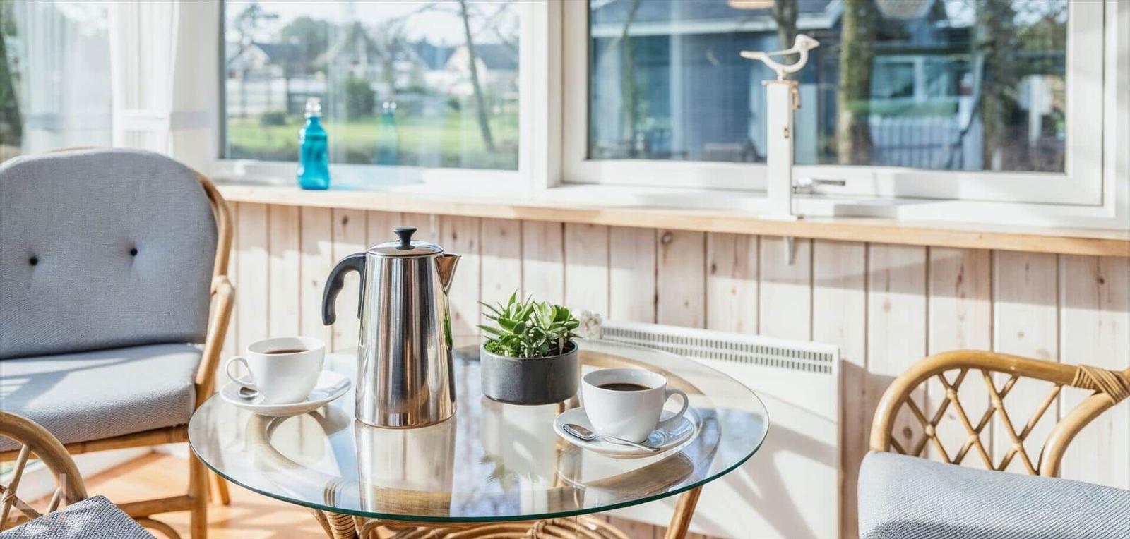 Glas-Tisch mit Kaffeekanne und Tassen vor Fenster mit Holzverkleidung.