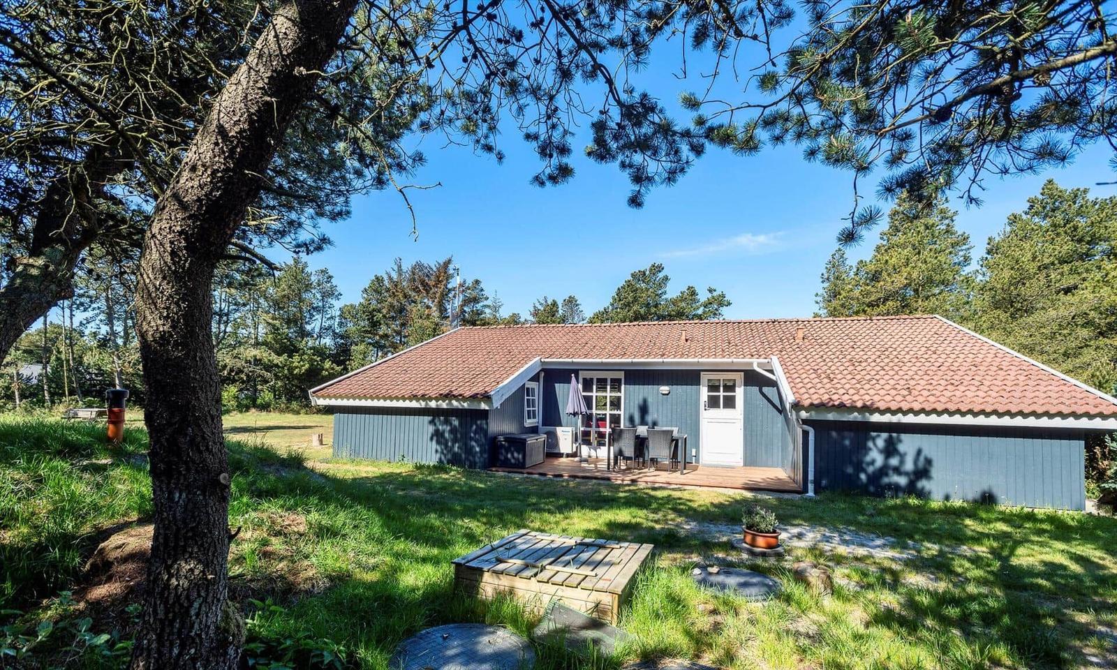 Ein Haus mit Terrasse und Garten unter Bäumen. Dachziegel und Holzverkleidung sichtbar.