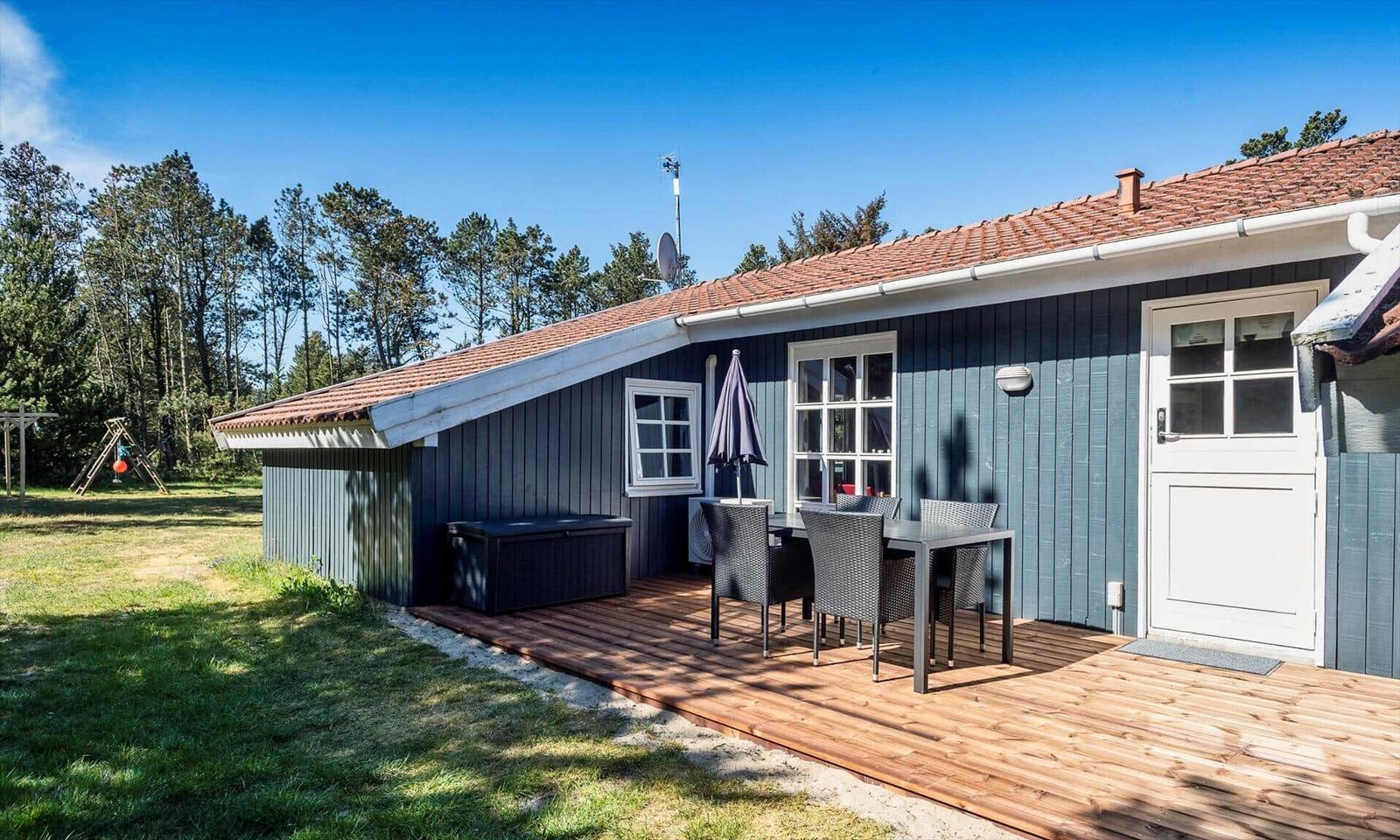 Haus mit Terrasse, Tisch, Stühlen und Sonnenschirm. Hintergrund: Wald und klarer Himmel.