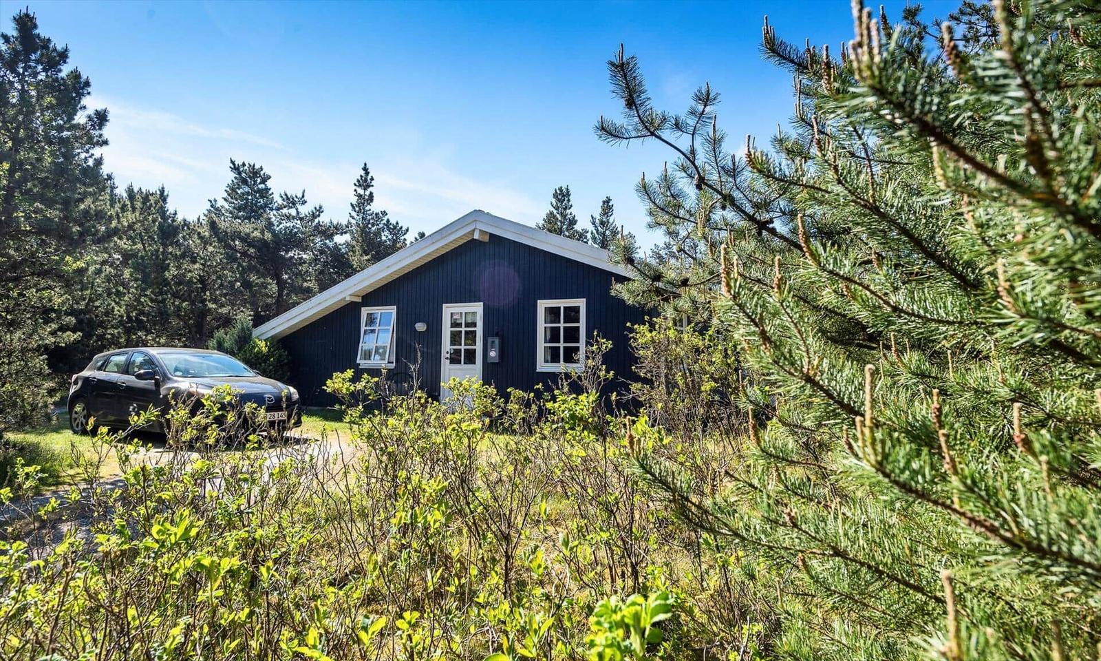 Schwarzes Holzhaus mit weißer Tür und Fenstern. Auto parkiert vor dem Haus. Umgeben von Bäumen und Gras.