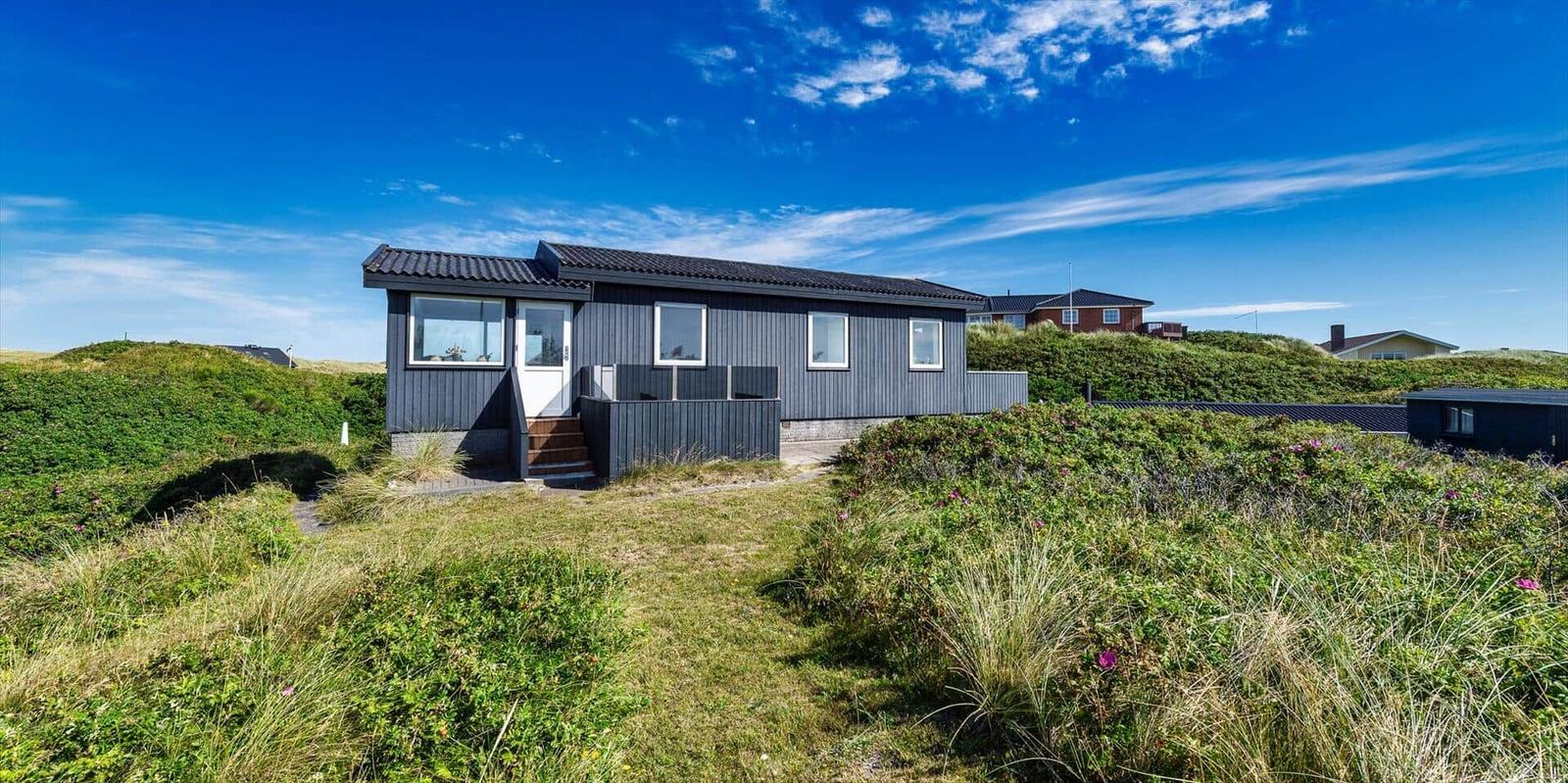 Ein graues Haus mit weißen Fenstern steht auf einer grünen Düne unter blauem Himmel.