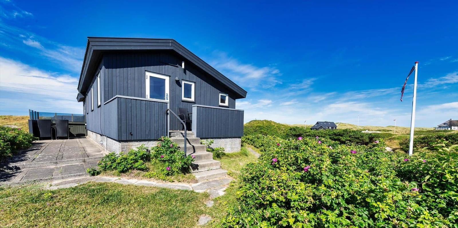 Moderne Strandhütte mit Terrasse und Garten. Blick auf Dünen und klaren Himmel.