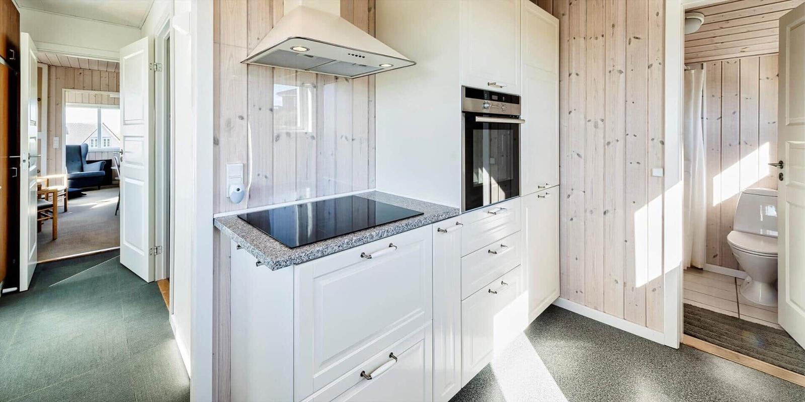Küche mit weißen Schränken, Induktionskochfeld und Backofen. Nebenraum mit Toilette und Wohnzimmer.