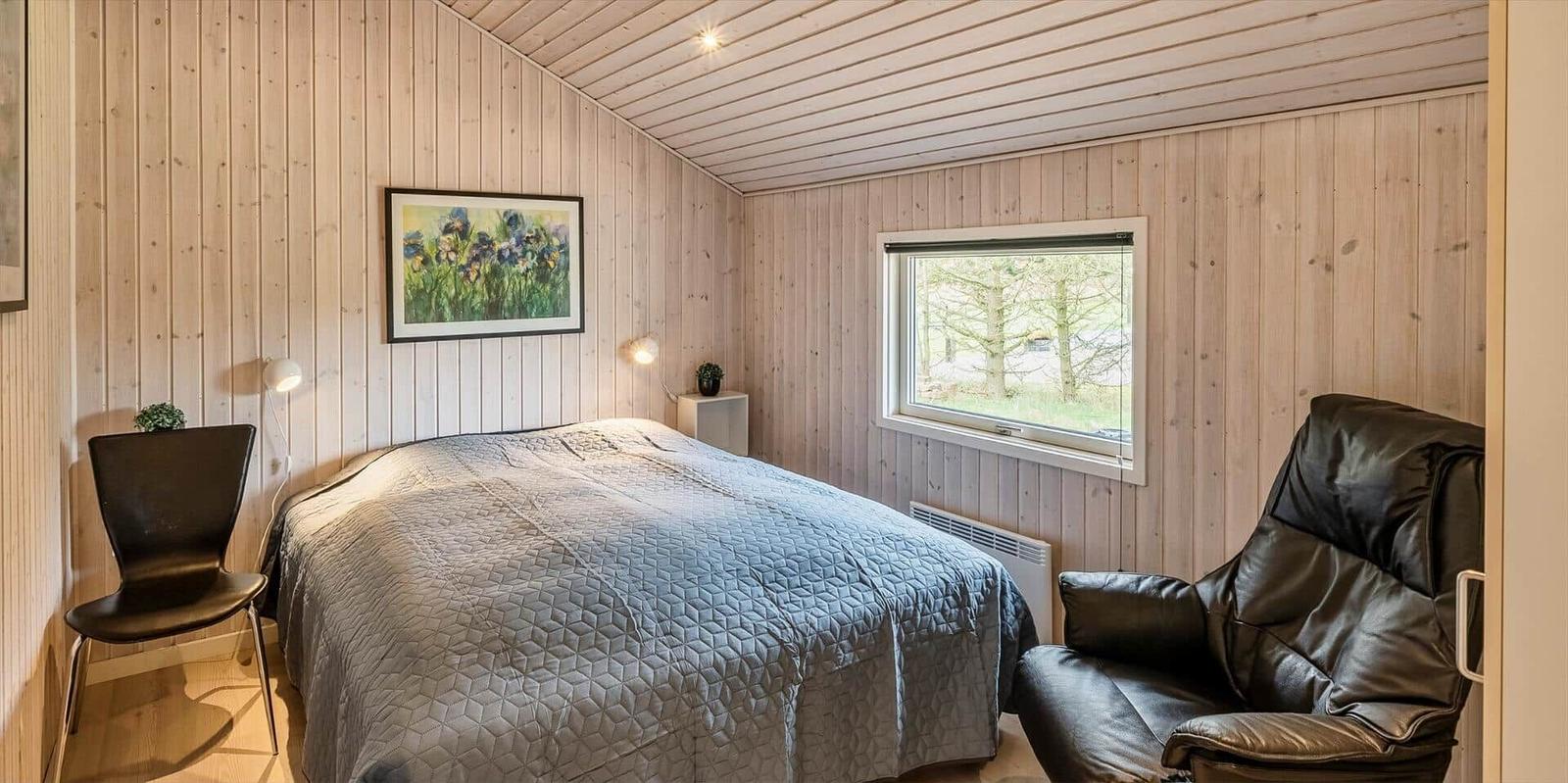Schlafzimmer mit Bett, Ledersessel und Holzwandverkleidung. Fenster mit Blick auf Bäume.