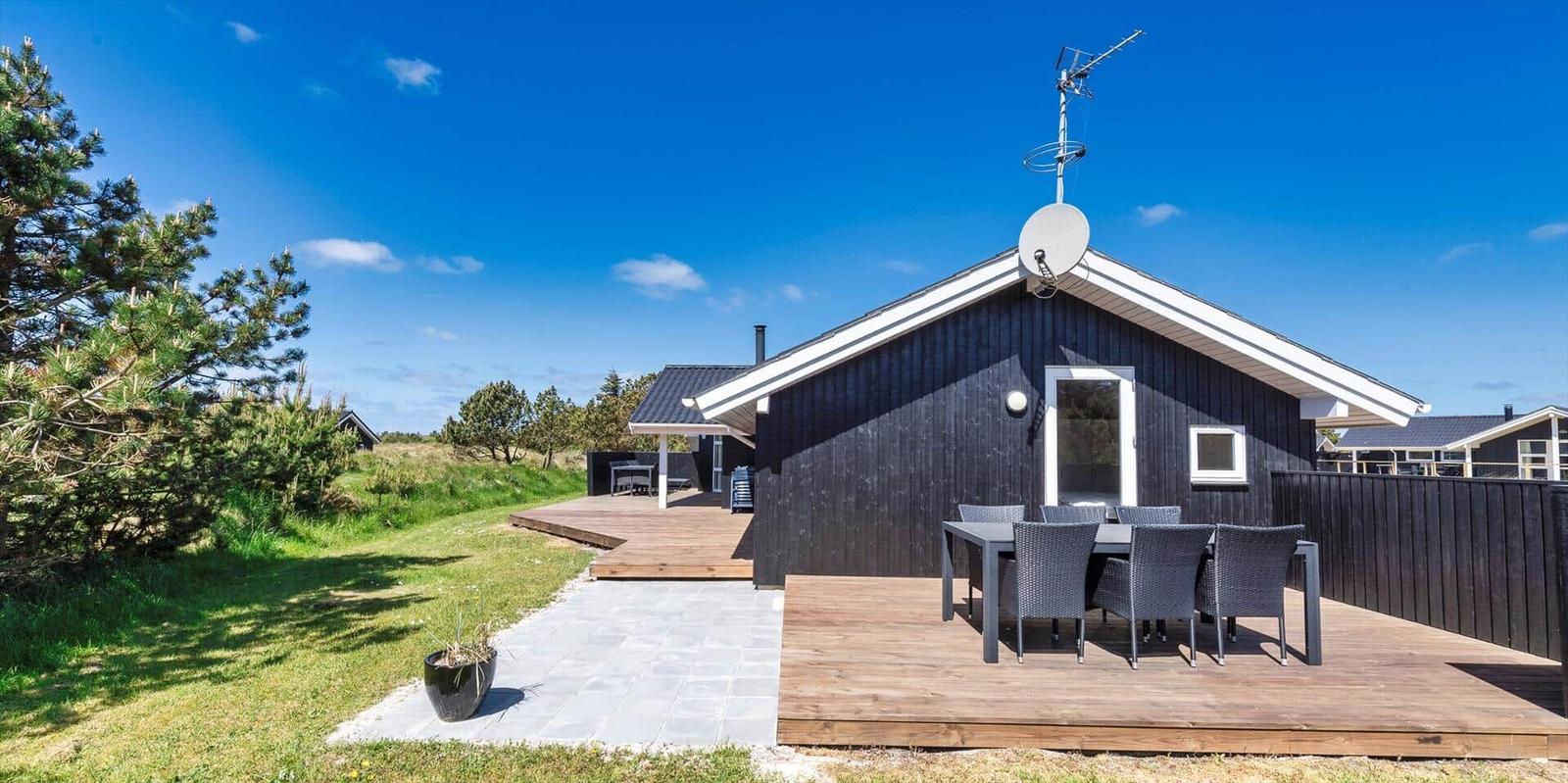 Schwarzes Holzhaus mit Terrasse und Garten. Satellitenschüssel und Außentisch sichtbar.