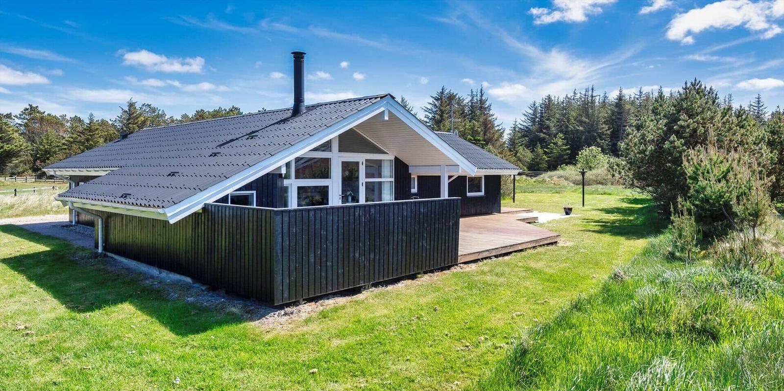 Moderne Holzhütte mit Terrasse und Garten. Umgeben von Bäumen unter blauem Himmel.