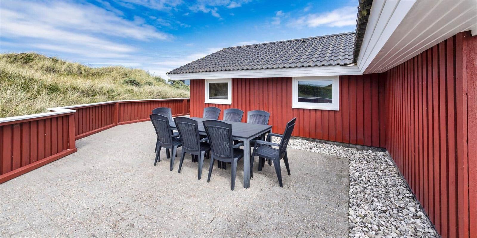 Roter Holzaussenbereich mit Tisch und Stühlen. Hintergrund: Dünen und blauer Himmel.