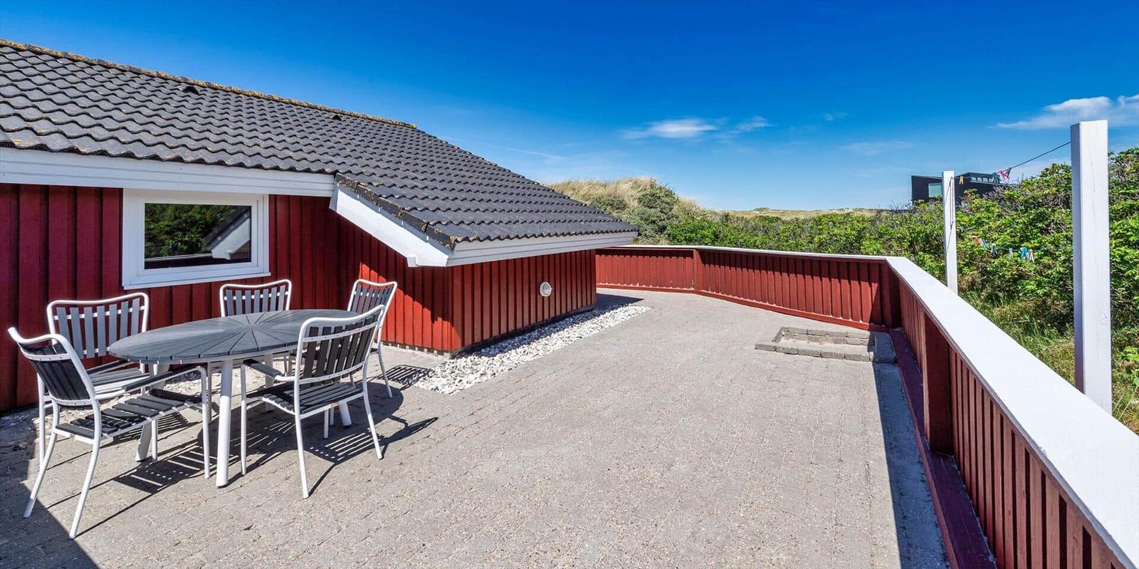 Roter Holzhaus mit Terrasse und Tisch. Blick auf grüne Dünen.