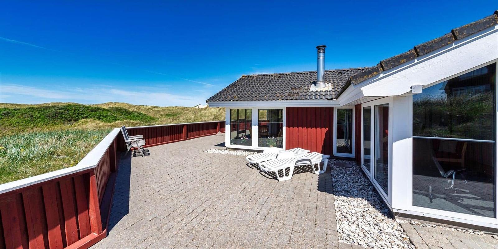 Rotes Ferienhaus mit Terrasse und Liegen. Blick auf Dünen unter blauem Himmel.