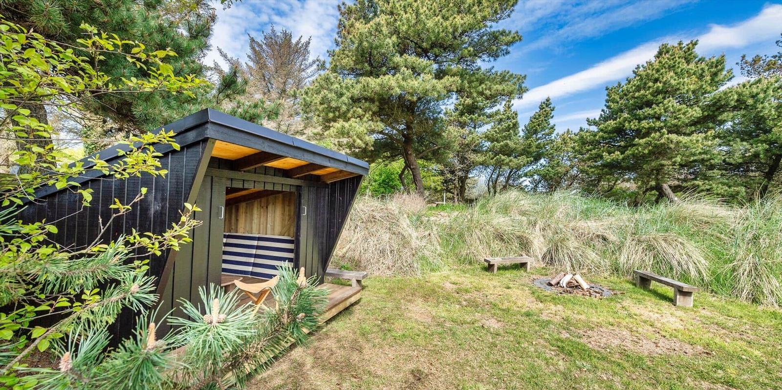 Ein kleines Holzhaus mit Sitzbank und Feuerstelle im Gras unter Bäumen.