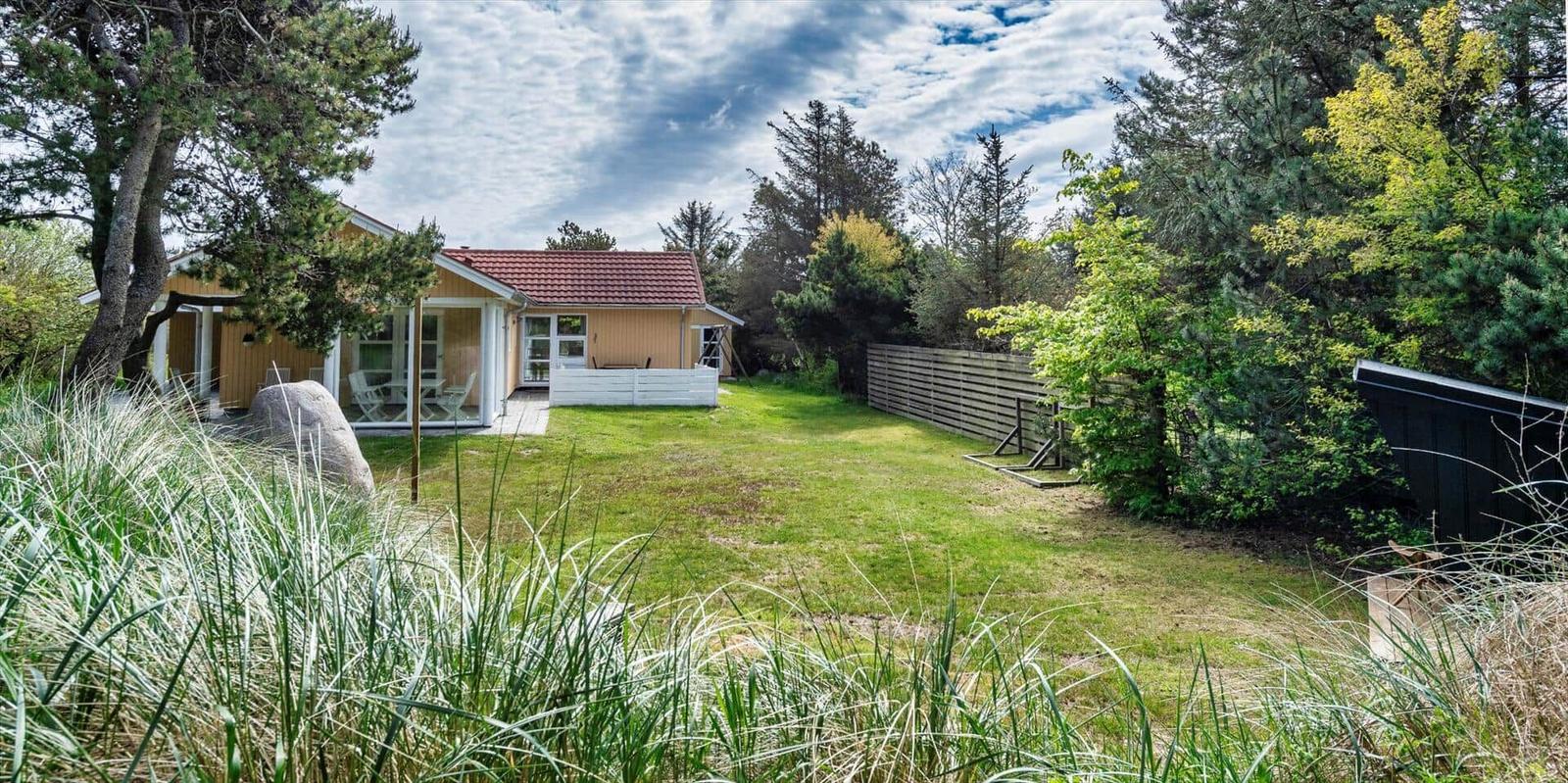 Haus mit Garten, Terrasse und Bäumen. Gras im Vordergrund.
