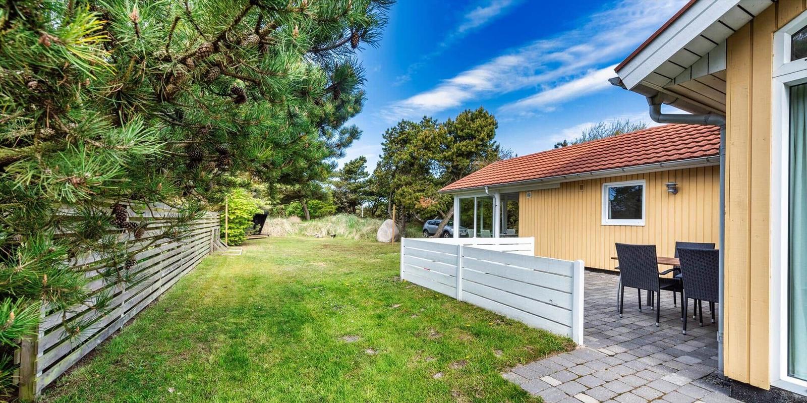 Haus mit Terrasse, Garten und Blick auf Bäume unter blauem Himmel.