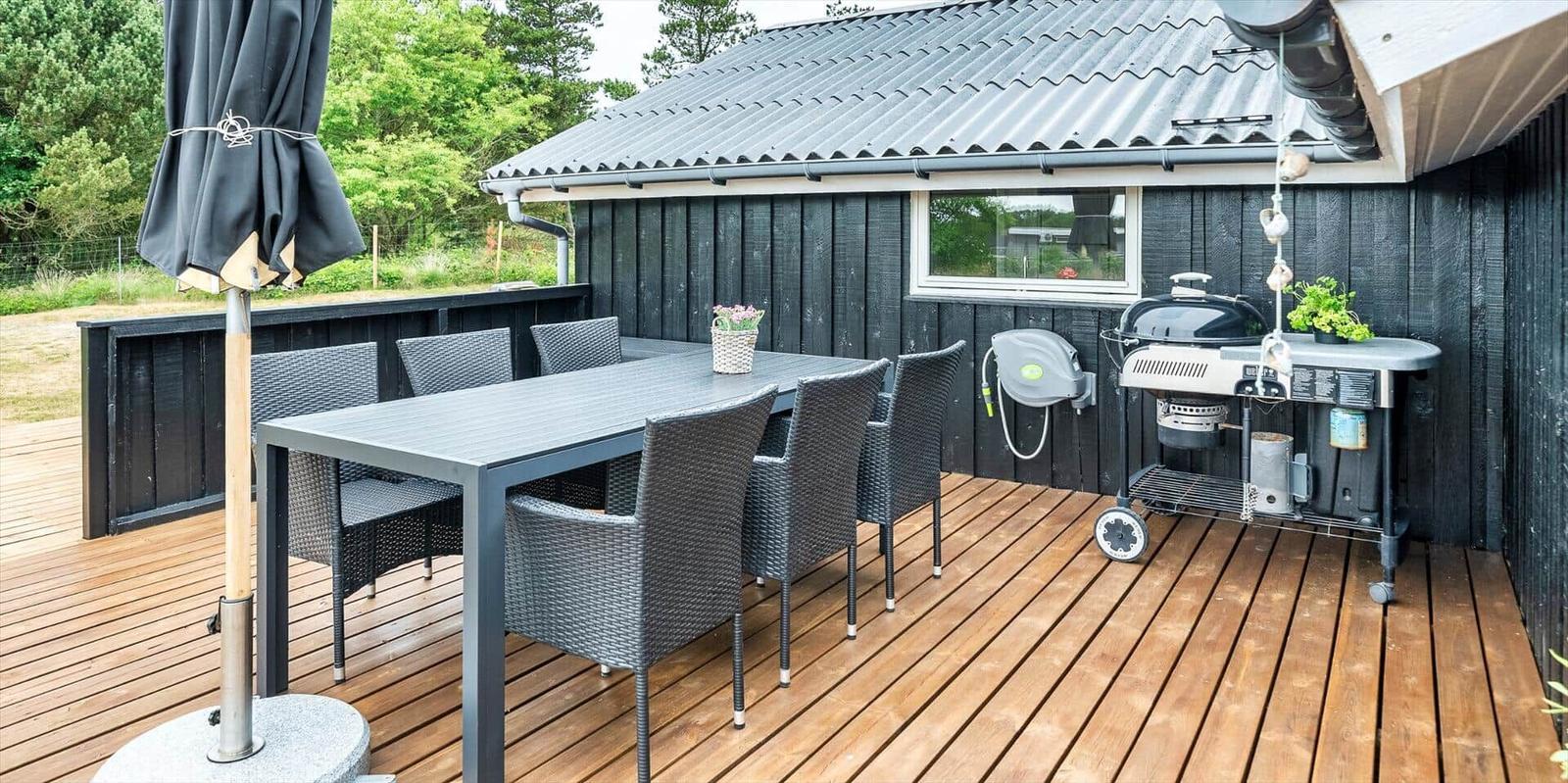 Terrasse mit Tisch, Stühlen und Grill. Hintergrund: dunkler Holzaufbau und Grün.