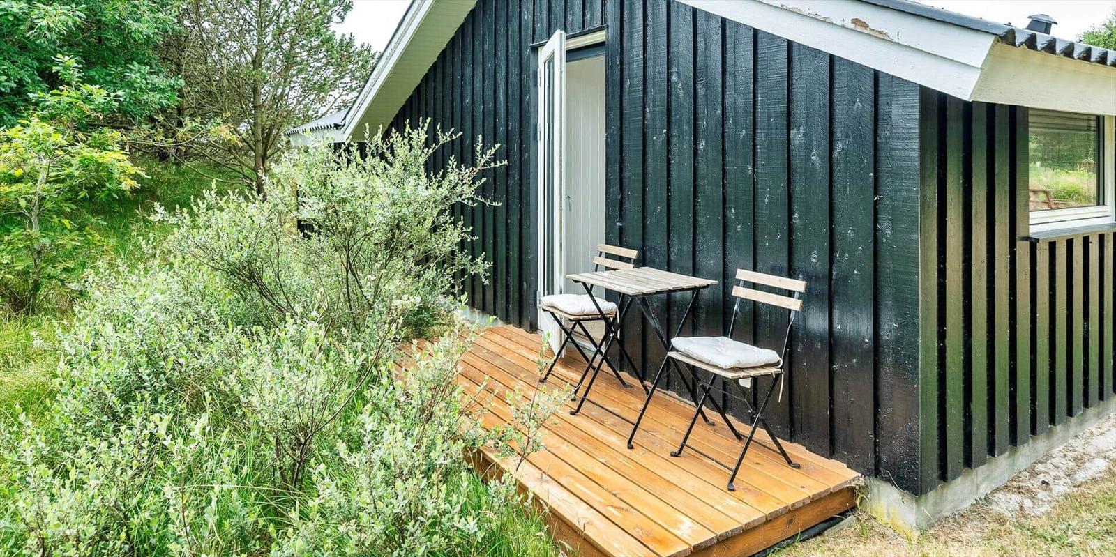 Hütte mit Holzterrasse, Tisch und zwei Stühlen. Umgeben von Grünflächen und Bäumen.
