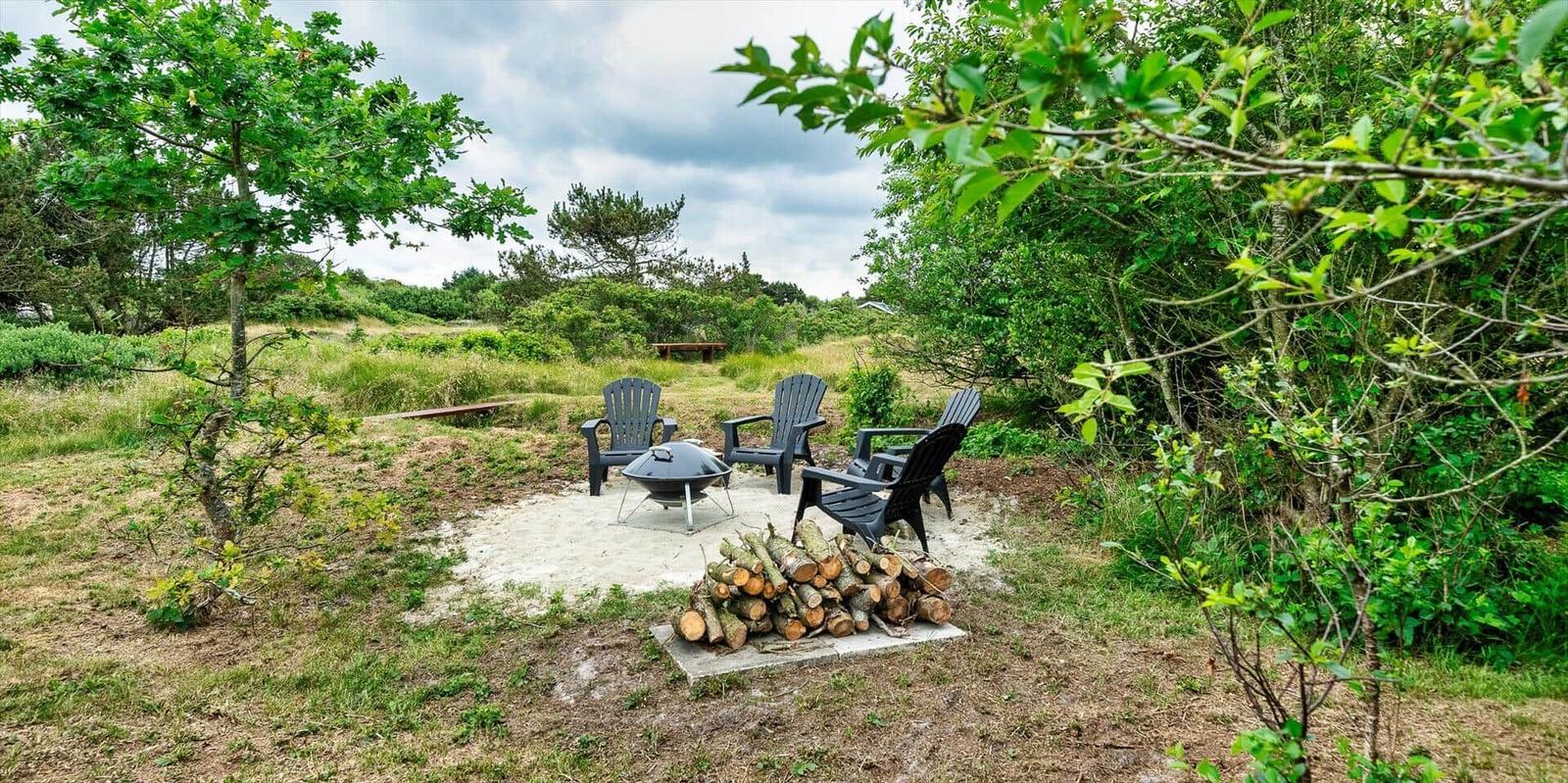 Außenbereich mit drei Stühlen, Grill und Holzstapel umgeben von Grünflächen.