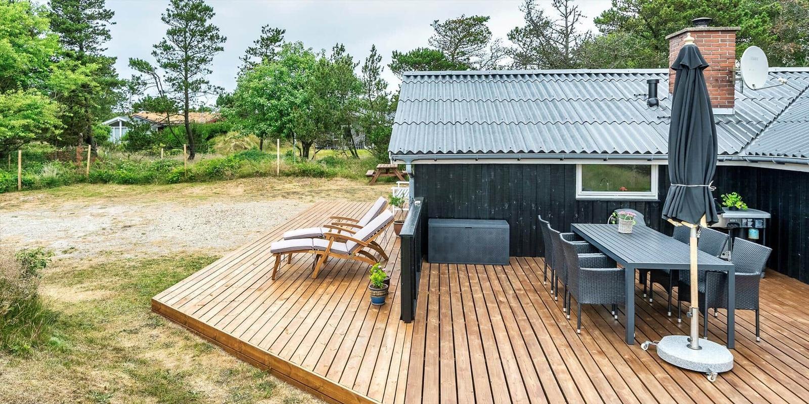 Holzterrasse mit Liegestühlen und Esstisch vor einem Haus mit grauem Dach.